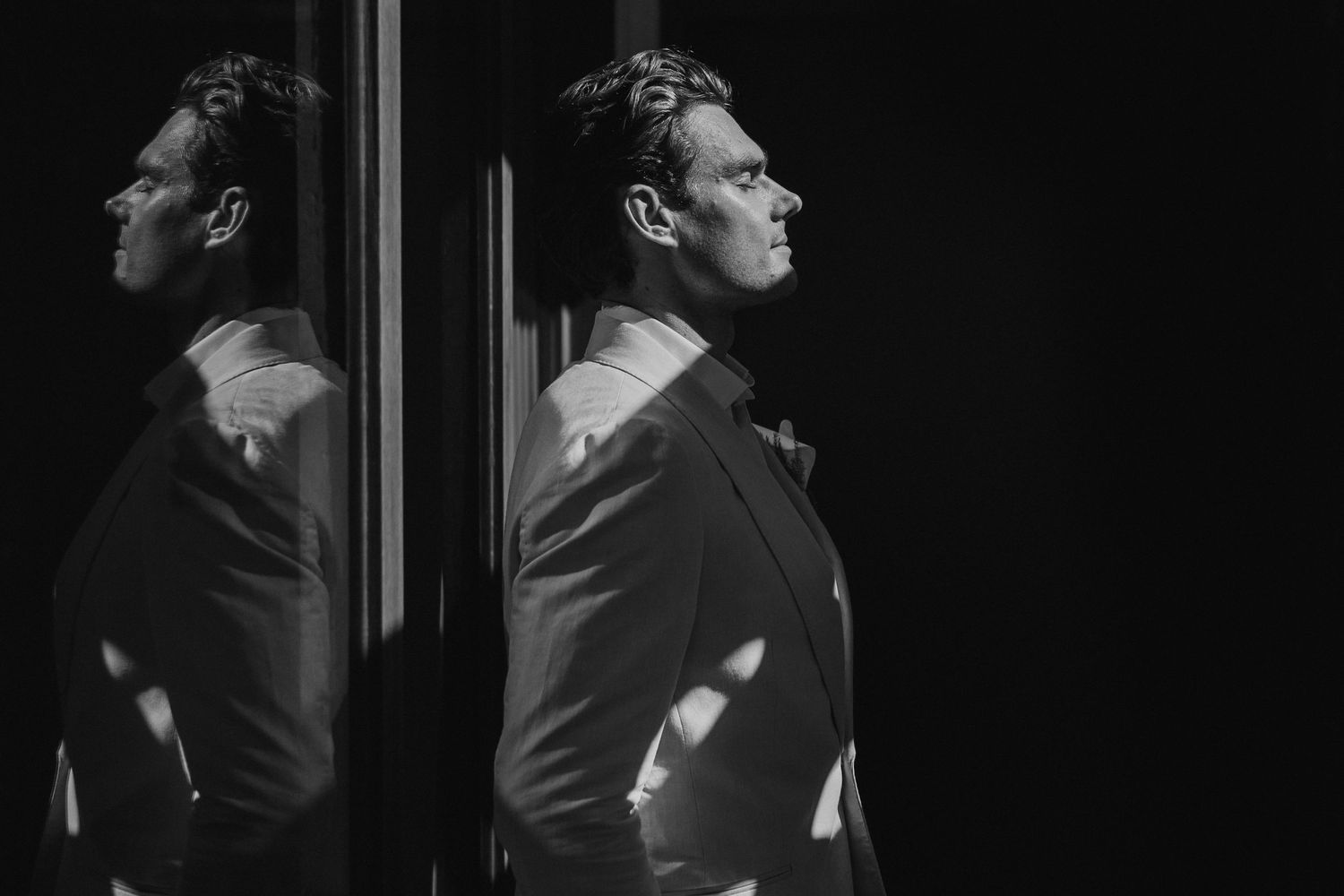 Elegant groom in a dark suit poses beside a mirror in a dramatic black and white portrait.