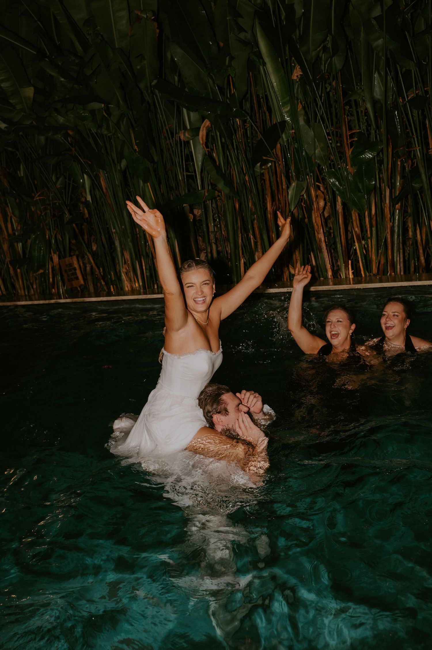 Bride in white dress celebrating in pool with friends at night during tropical wedding after-party.