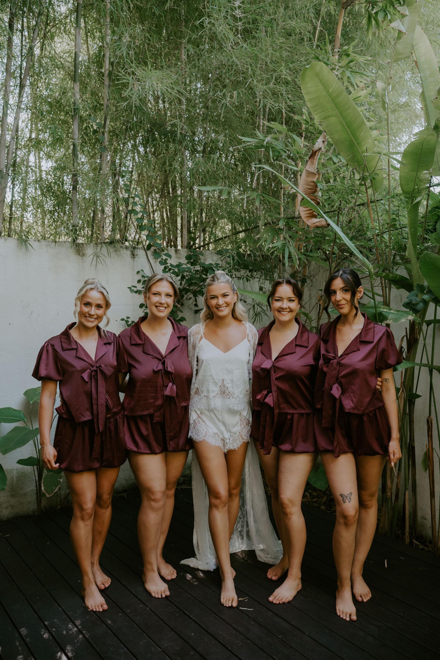 Bride in white lace robe poses with four bridesmaids wearing matching burgundy satin shorts sets outdoors.