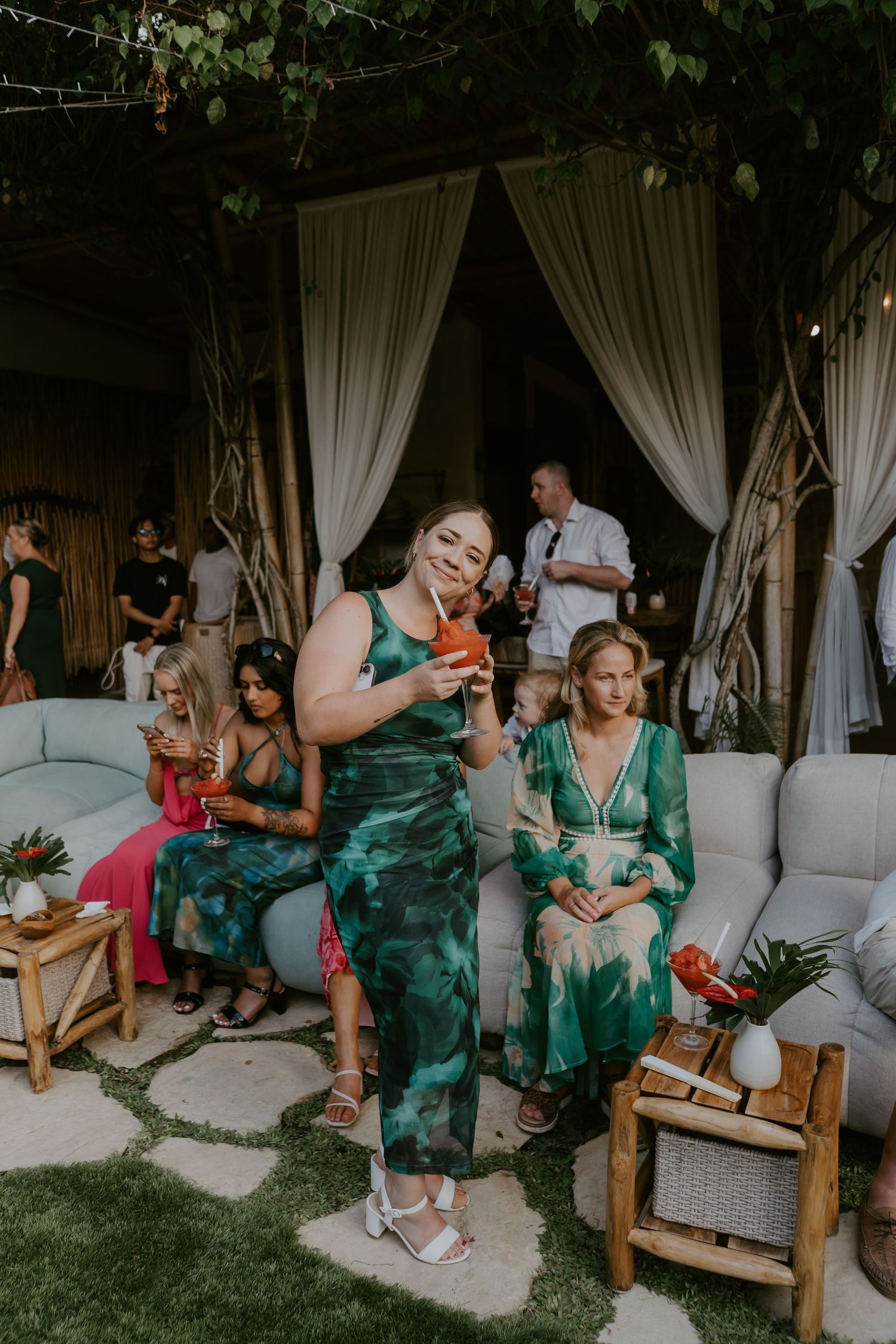 Wedding guest in green tie-dye dress enjoys cocktail hour at tropical outdoor garden reception.