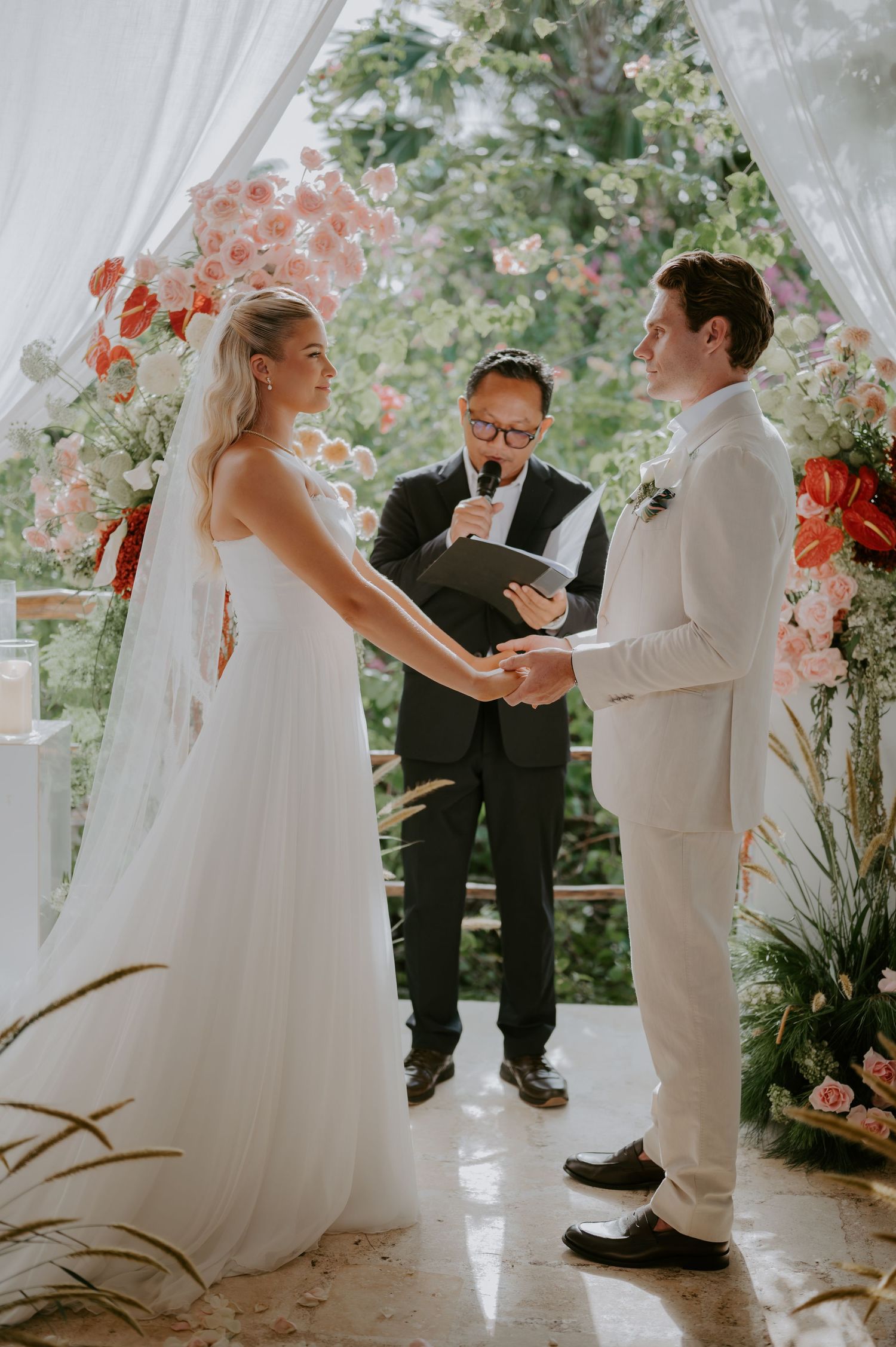 Bride and groom exchange vows at outdoor wedding ceremony with officiant and pink floral arch backdrop.