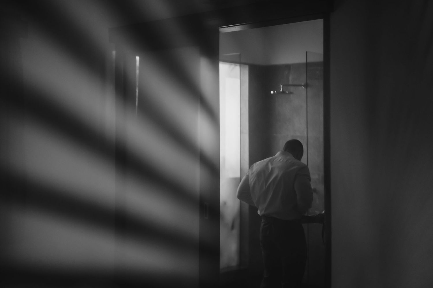 Man in white shirt stands in a softly lit bathroom doorway, captured in moody black and white photography.
