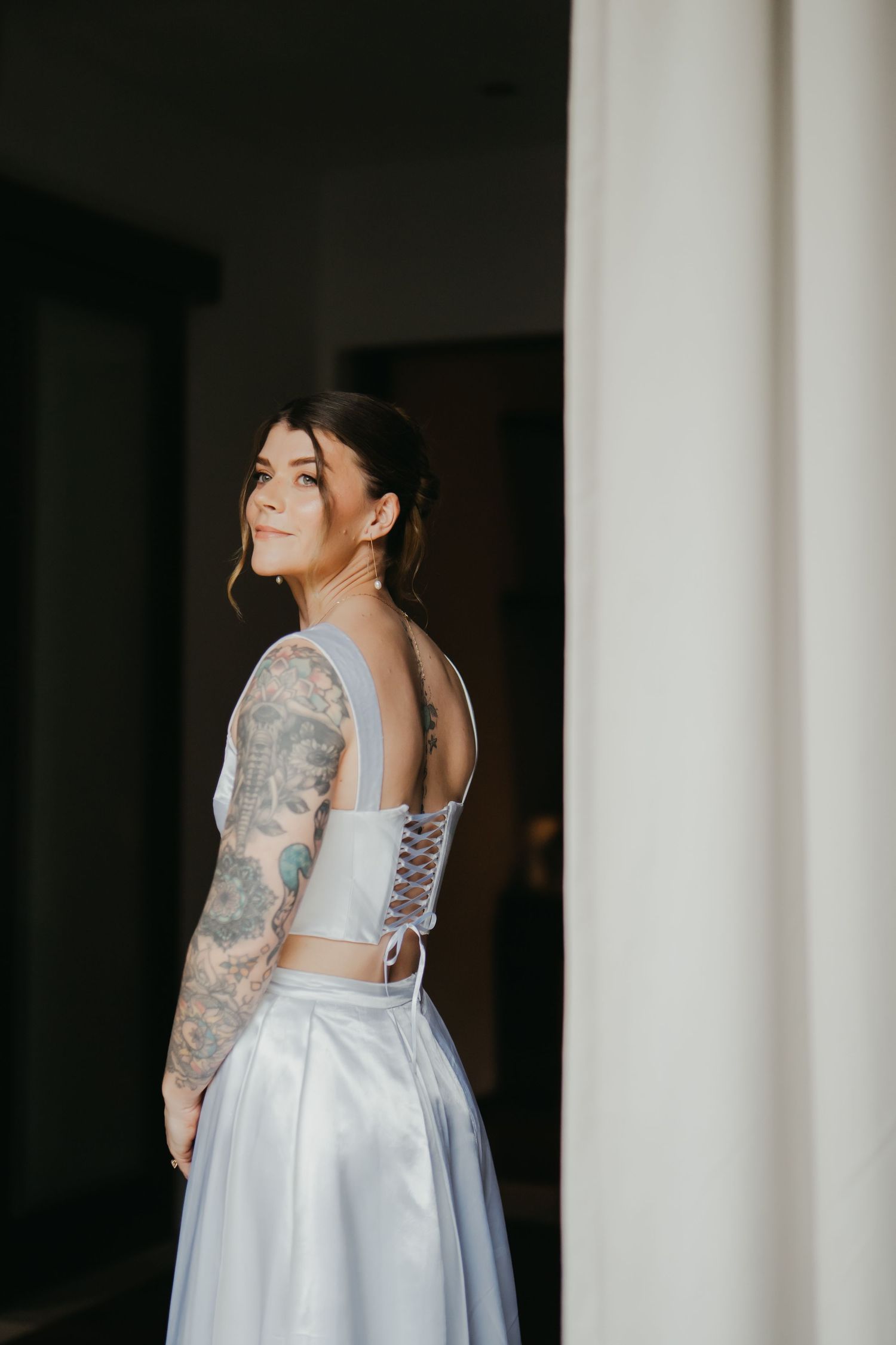 Bride in blue corset wedding gown with sleeve tattoos posing near white column.