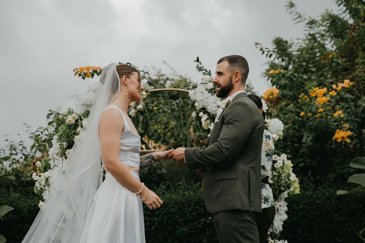 Bride and groom holding hands at outdoor ceremony beneath white and yellow floral arch.