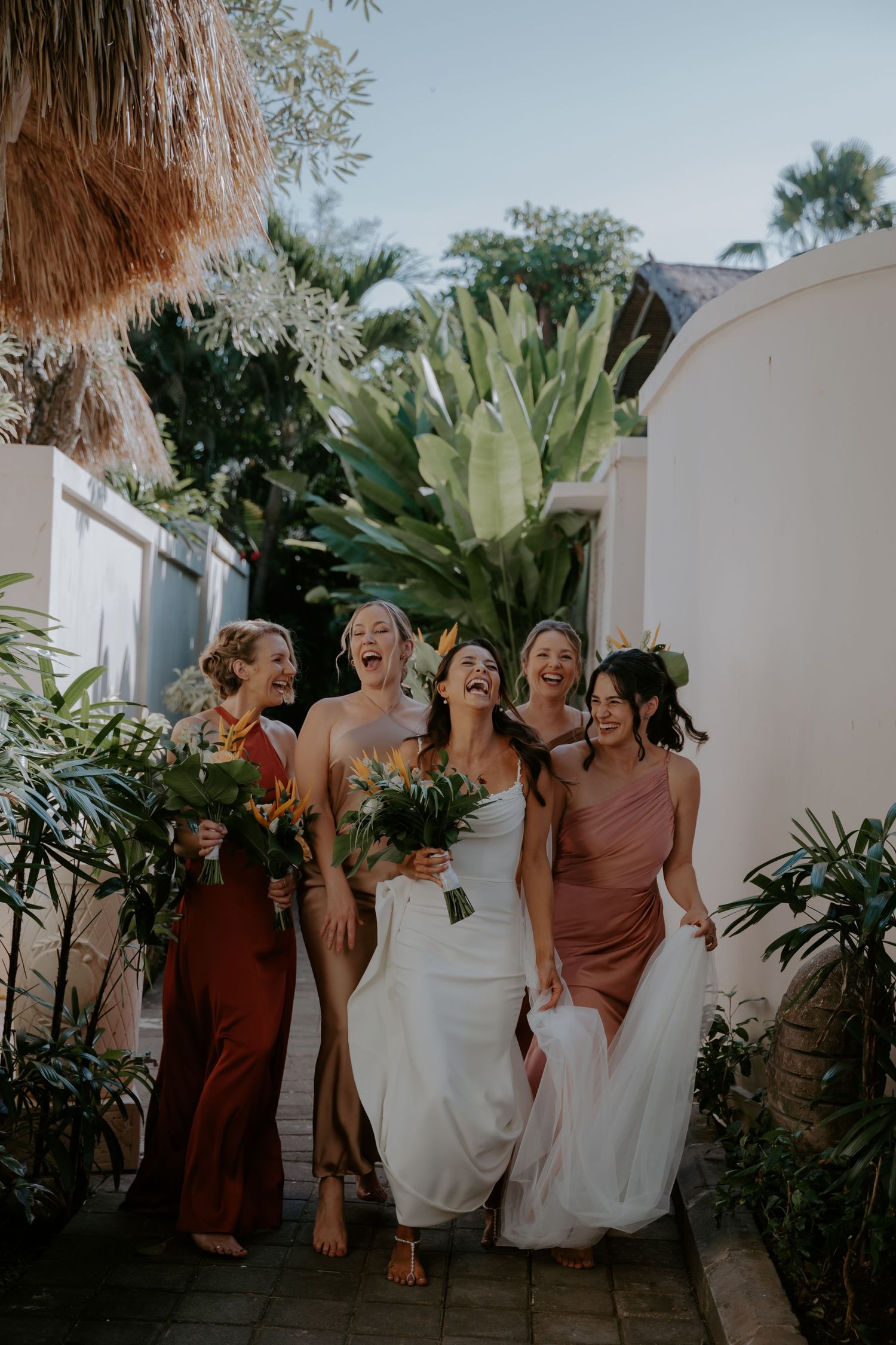 A group in brown and white dresses laugh and dance together in a tropical garden setting at a celebration.
