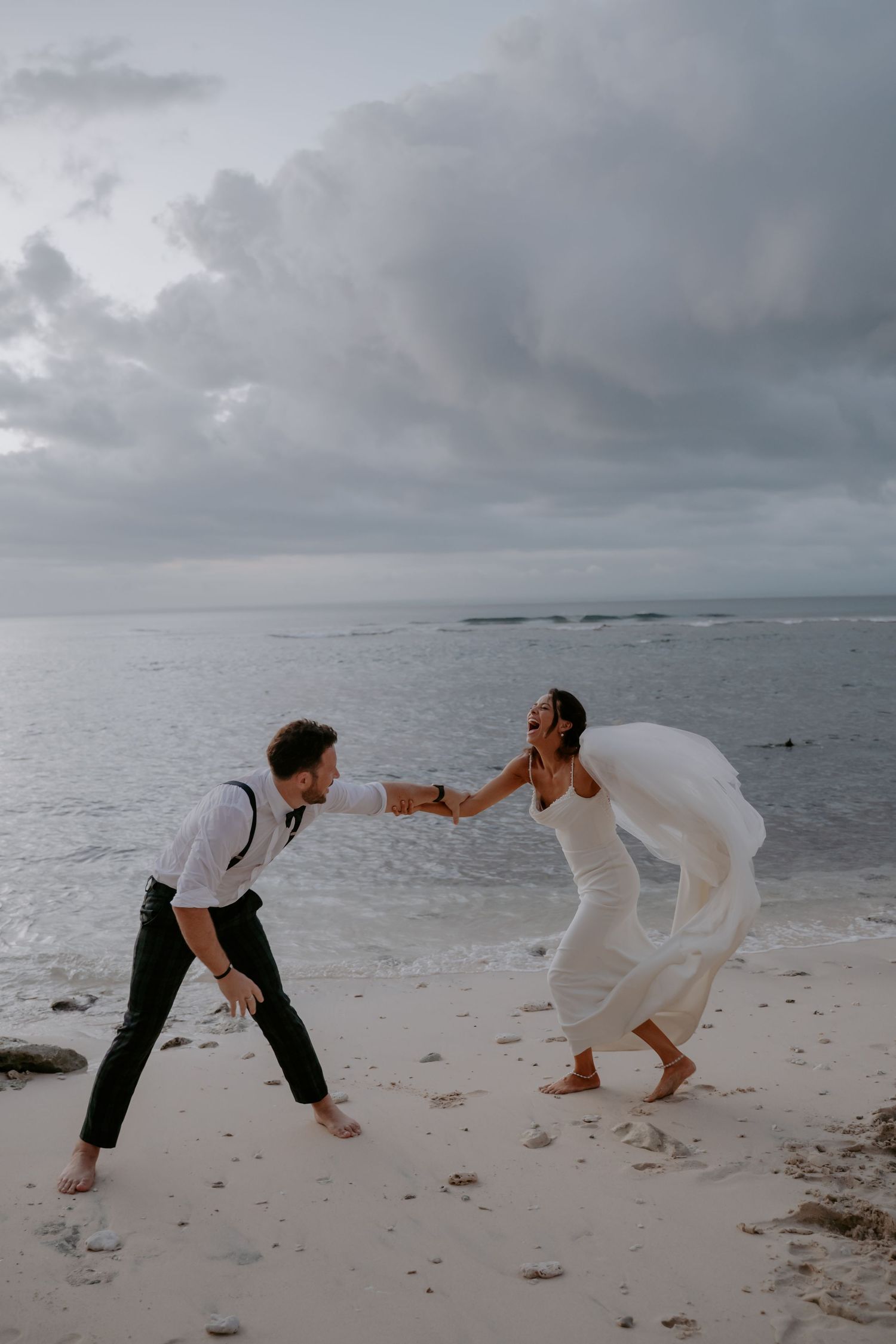 A couple in wedding attire playfully dance together on a cloudy beach with ocean waves in the background.