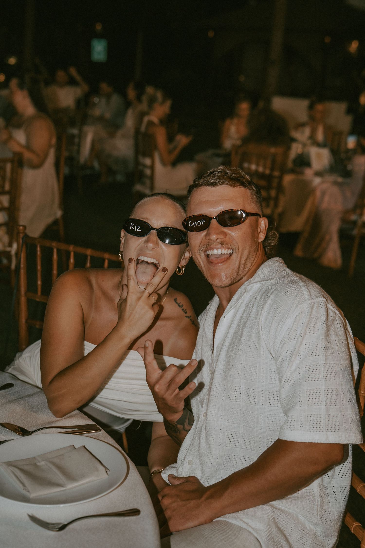 Two people wearing sunglasses laugh together at a dimly lit indoor event while sitting at a table.