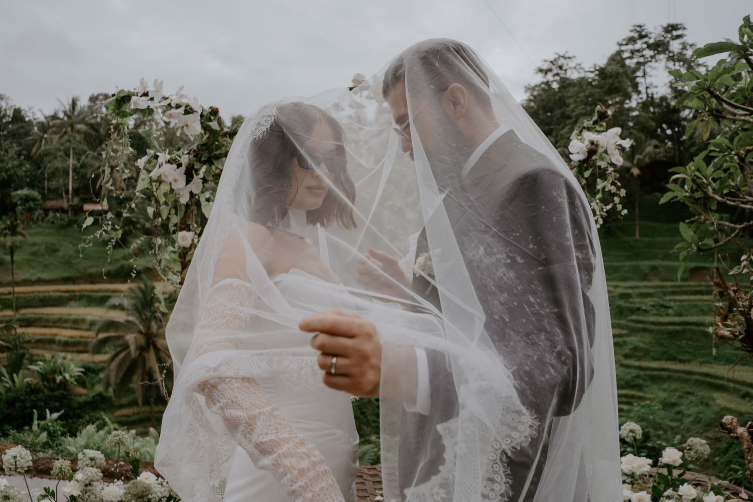 A romantic wedding couple embraces under a flowing veil in a lush garden setting.