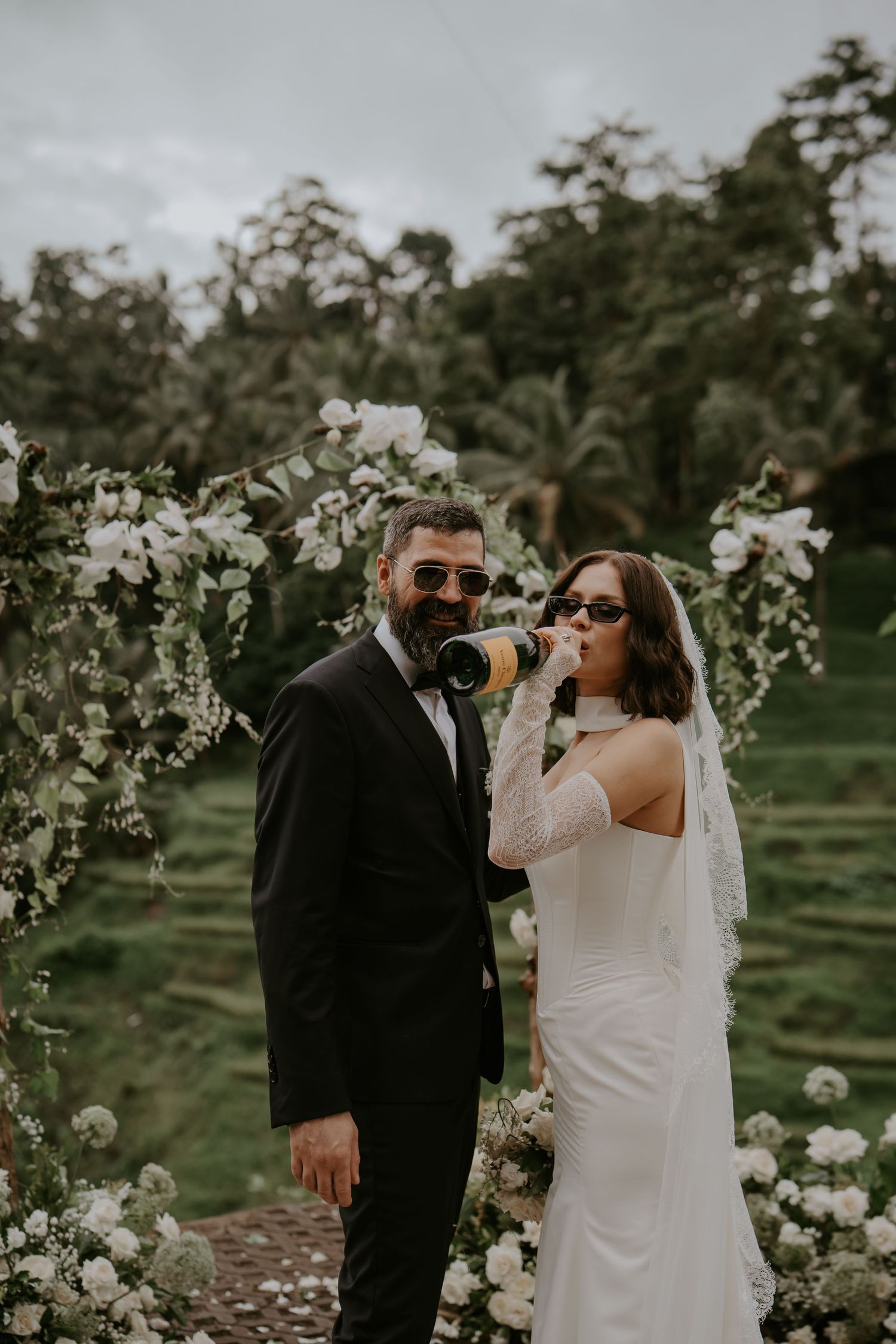 Newlyweds in sunglasses drinking beer together in front of floral arch at outdoor garden wedding ceremony.
