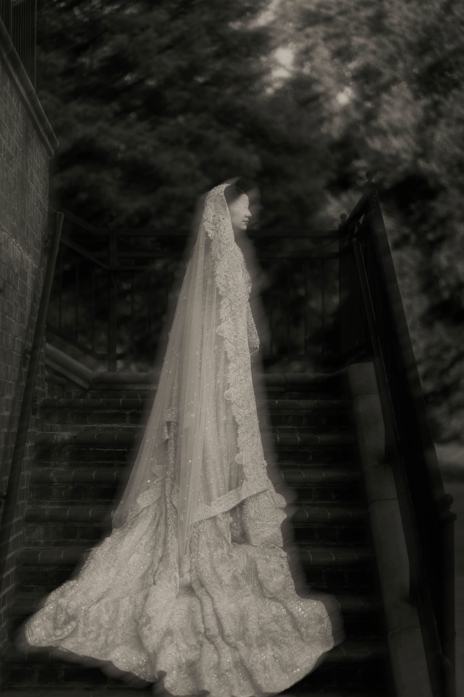 Black and white photo of a bride in a long lace wedding gown and veil standing on outdoor stone steps.