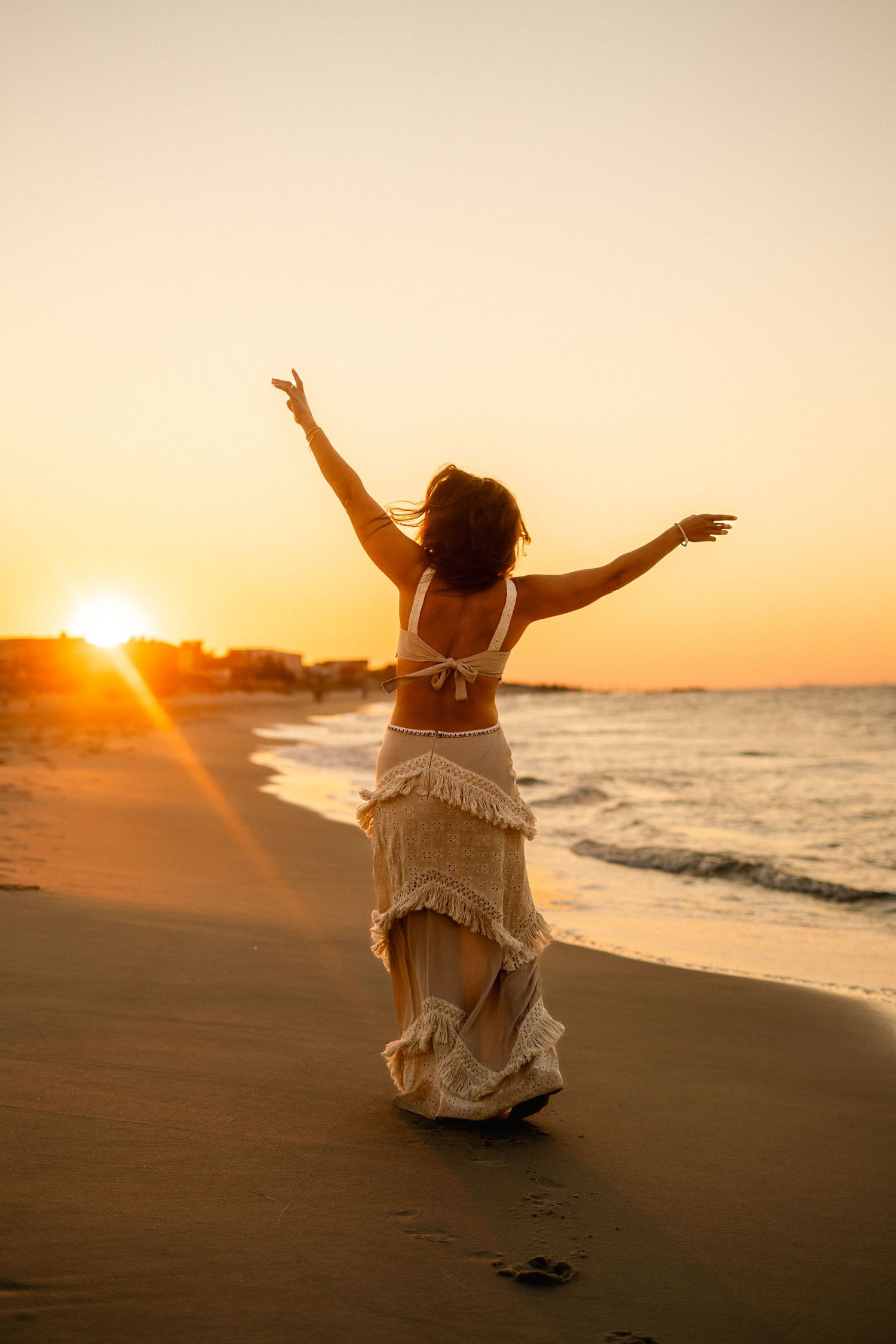 Woman in white fringe dress with arms raised celebrating on beach at golden sunset.