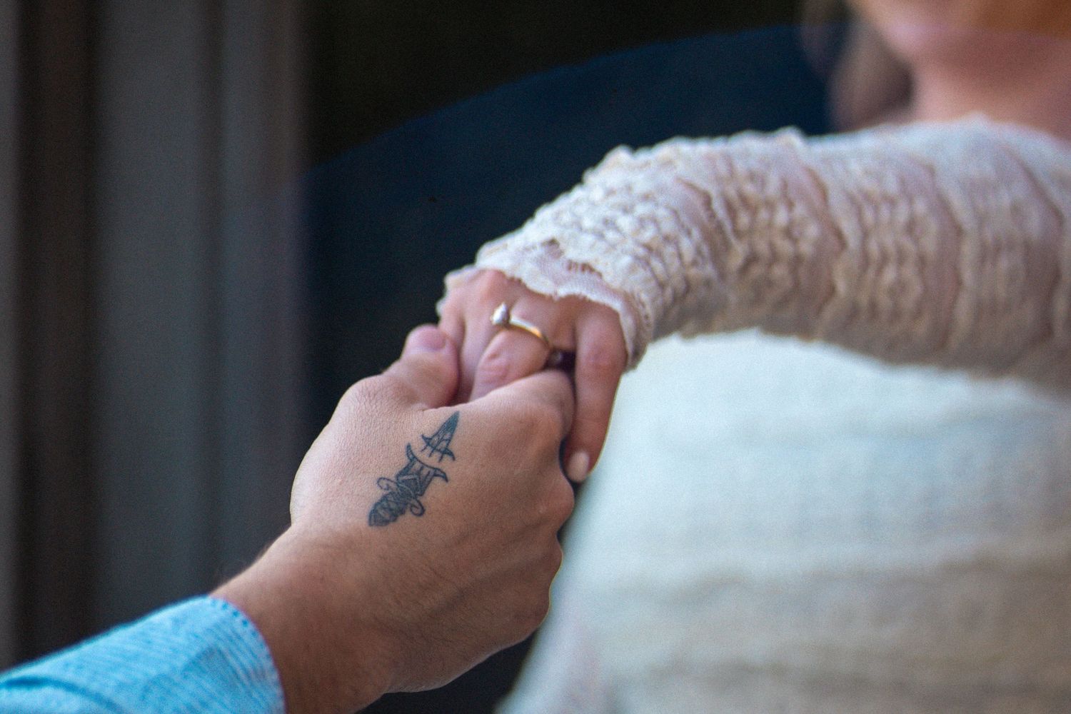 Close-up of two people holding hands, one with a cross tattoo, wearing wedding rings in a romantic ceremony.
