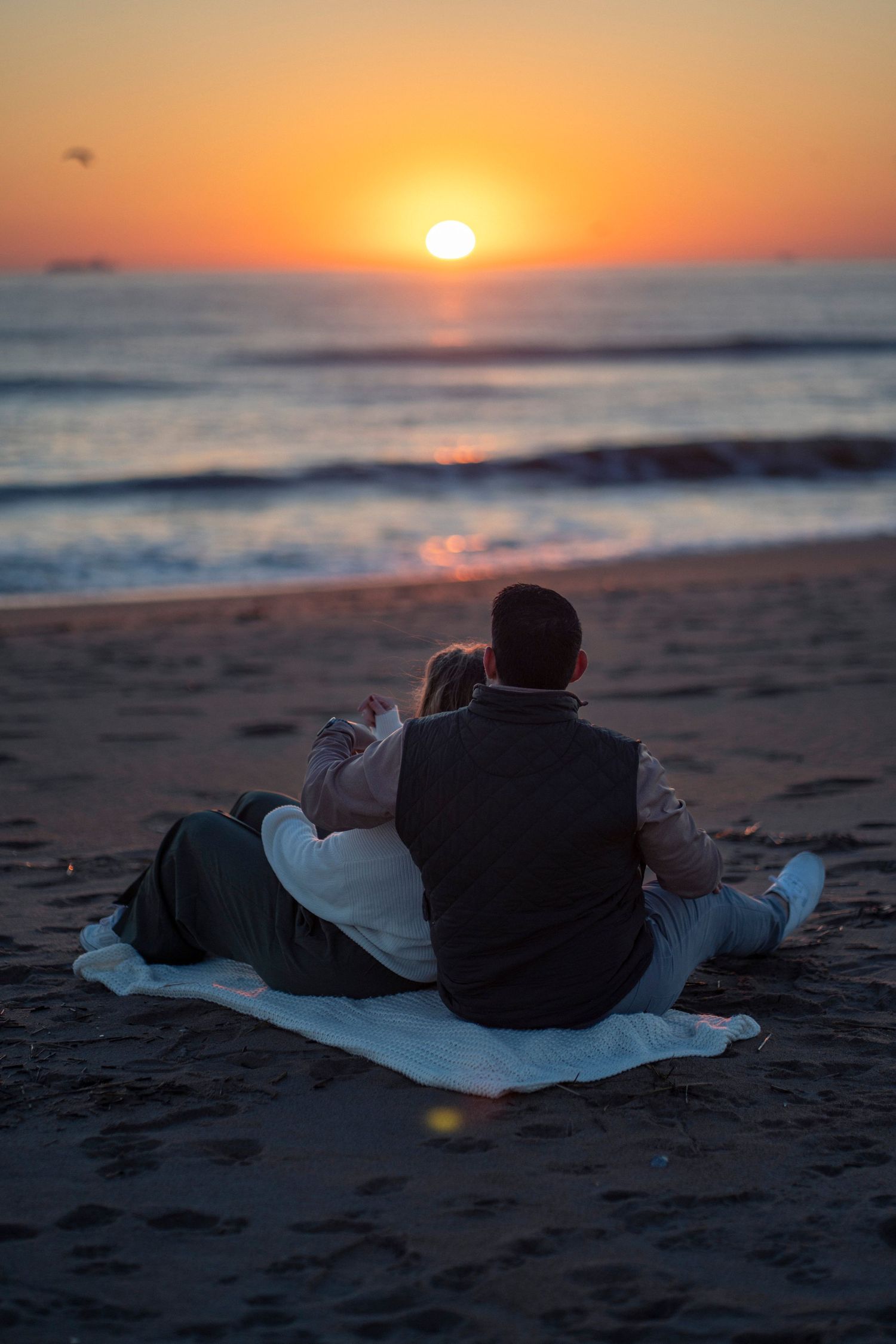 Couple sitting on a blanket on the beach watching a beautiful orange sunset over the ocean.