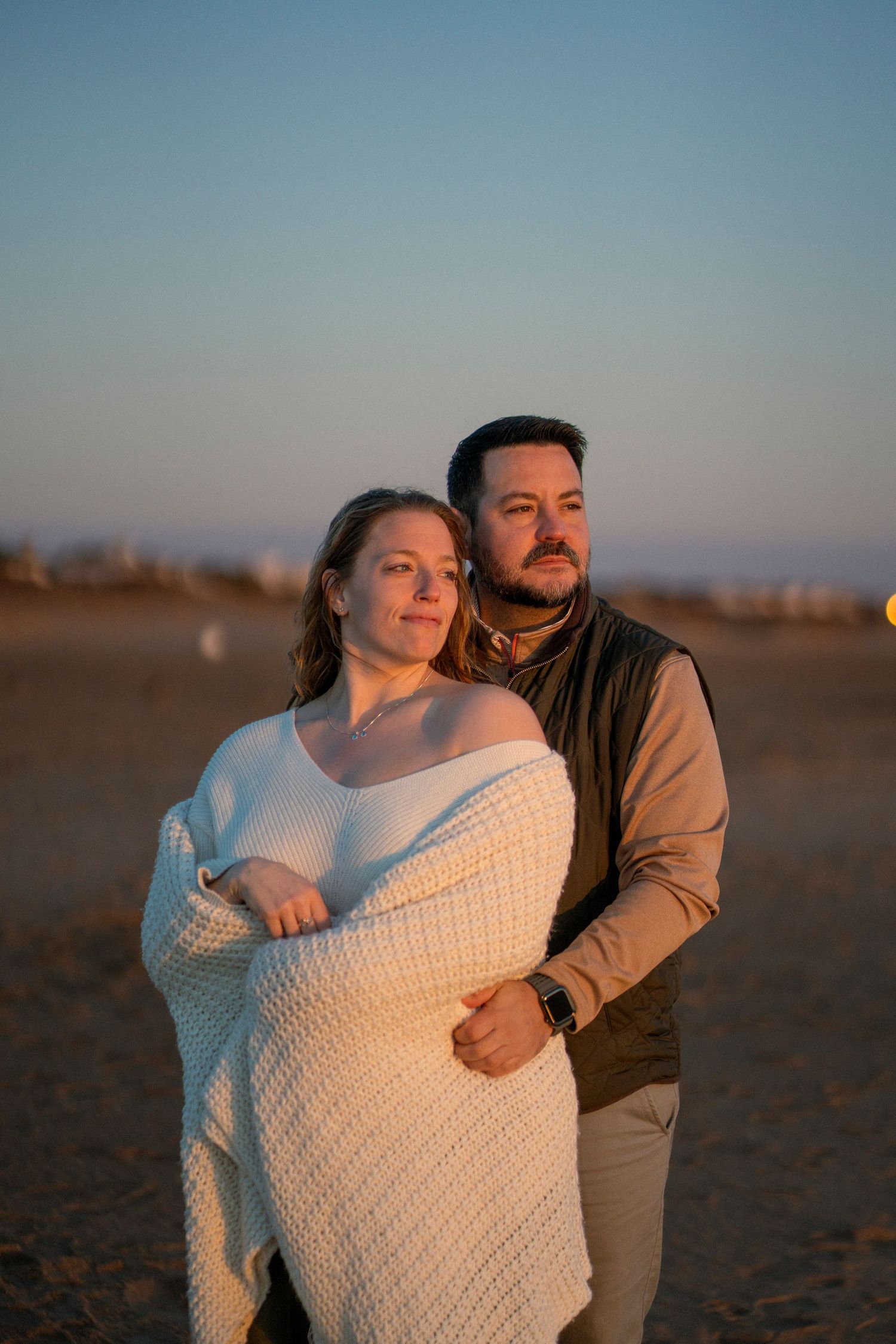 A man and woman wrapped in a blanket looking at the sunrise on the beach.