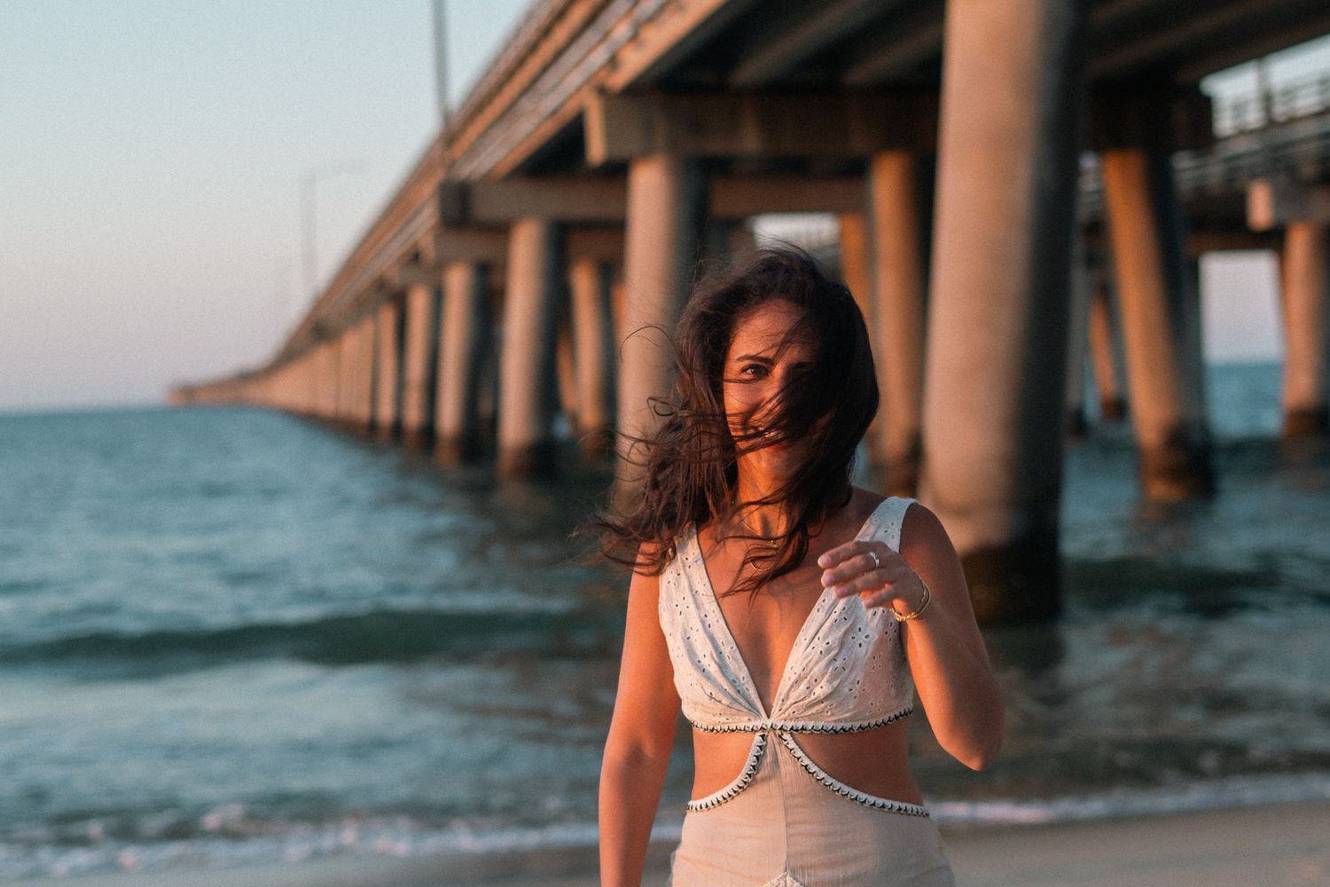 Woman in white bikini top poses on sandy beach near large bridge pillars at sunset with wind-blown hair.