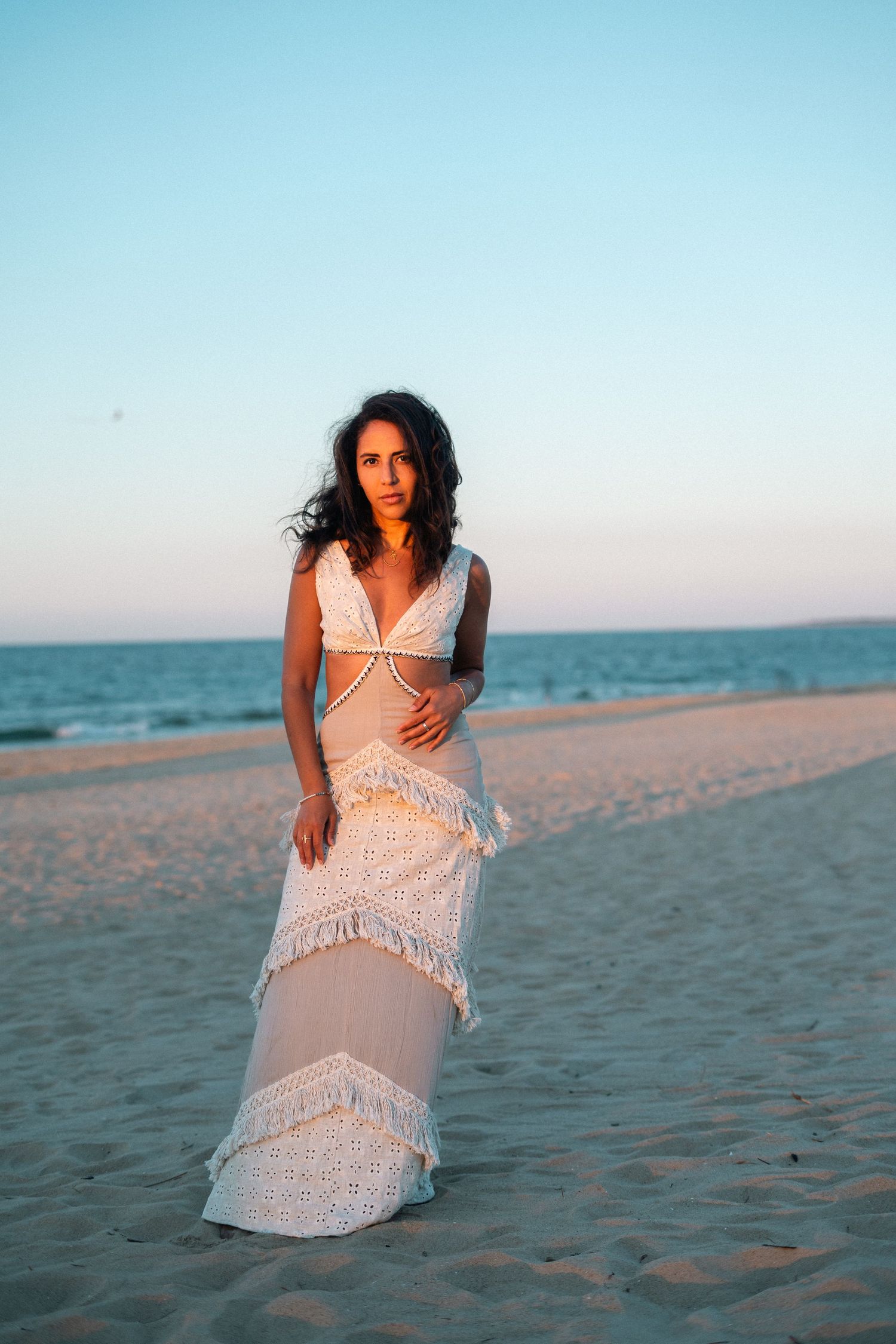 Woman in white lace fringe maxi dress posing on sandy beach at golden hour sunset.