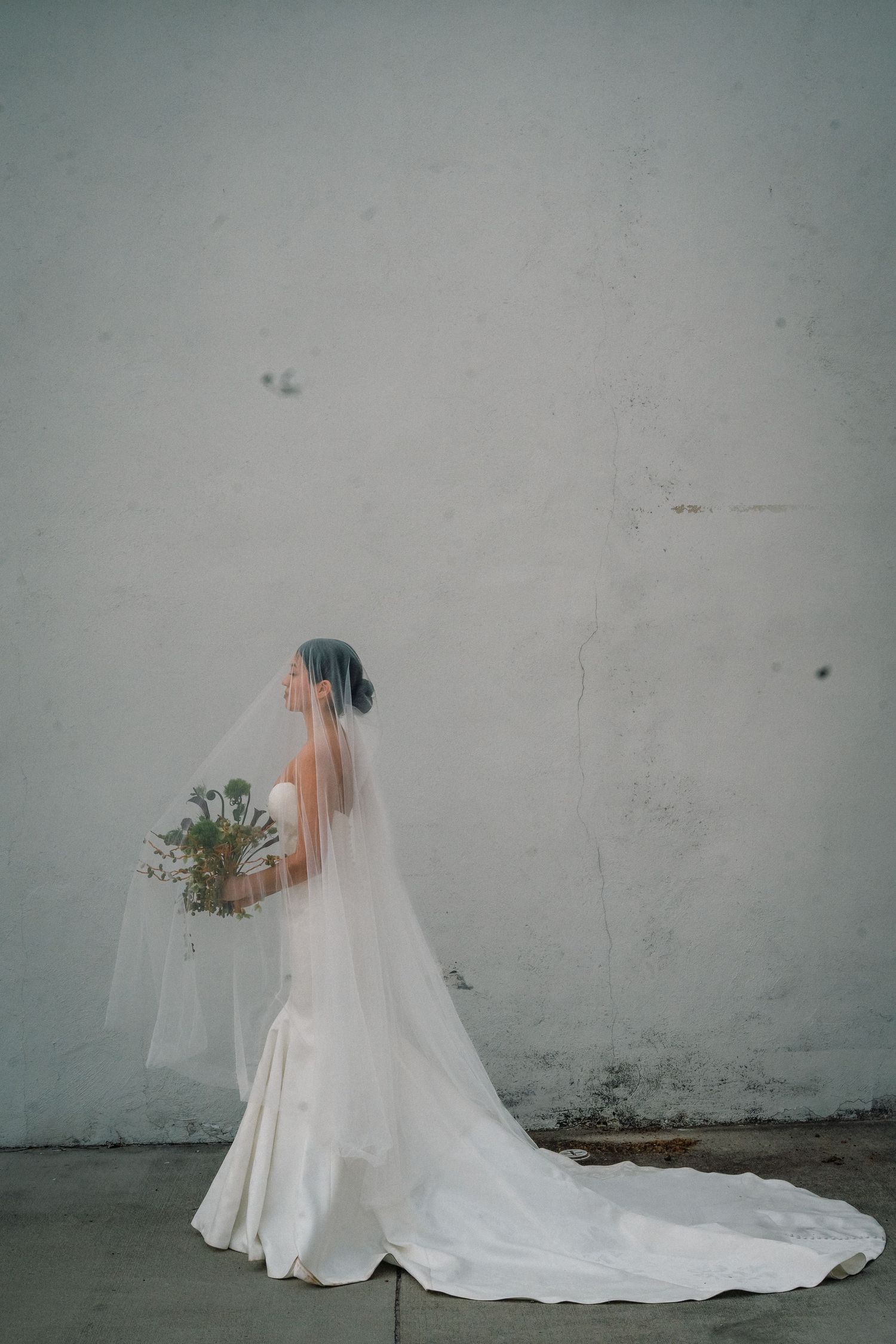 A bride looking off camera holding a bouquet with a long duster veil covering her face.