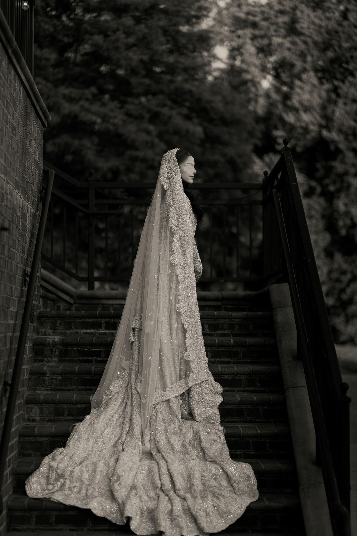 Black and white photo of a bride in a lace gown and long veil standing on stone steps outdoors.