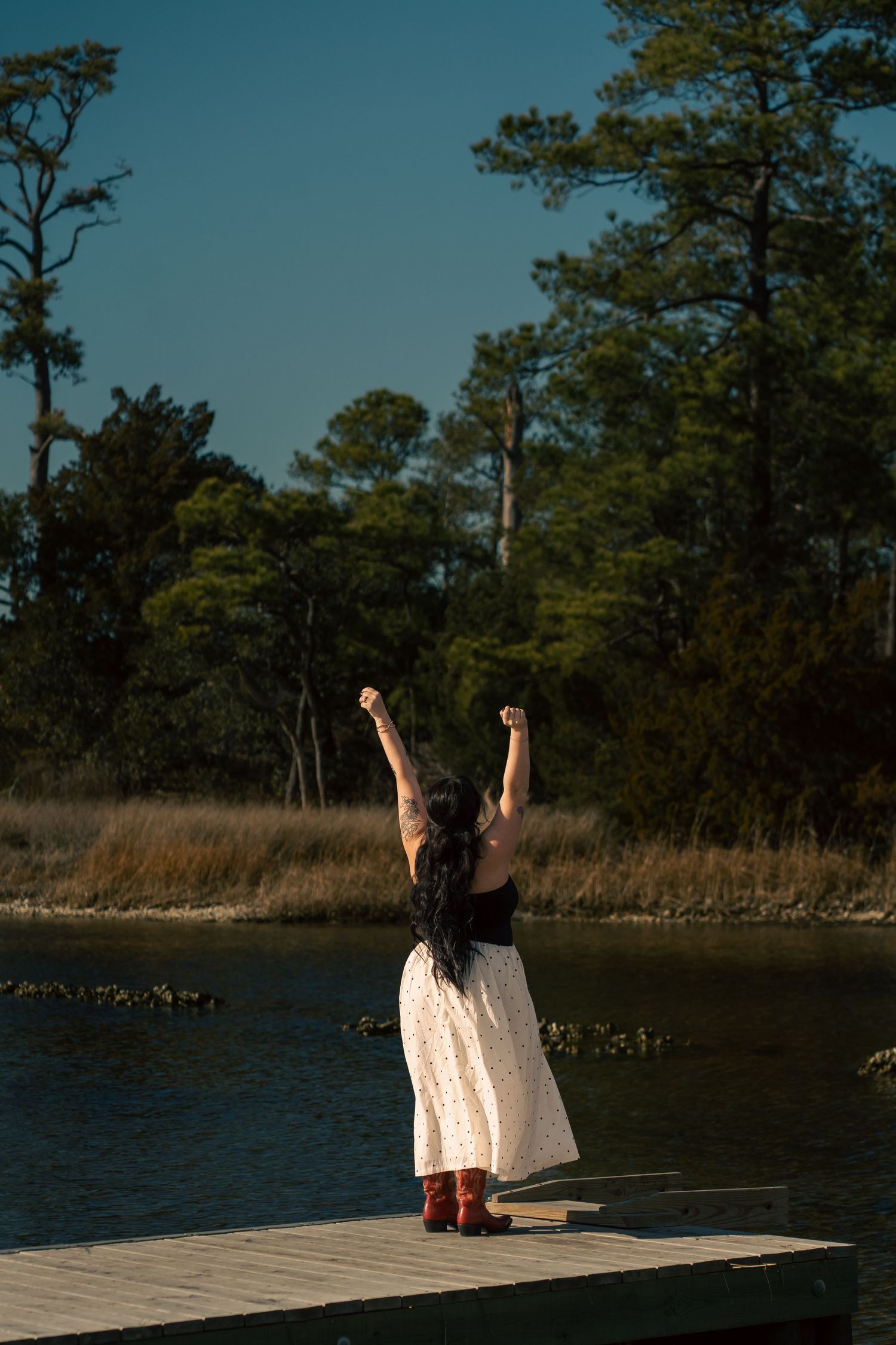 A woman in a white skirt and red cowboy boots raises her fists triumphantly on a dock by a river.