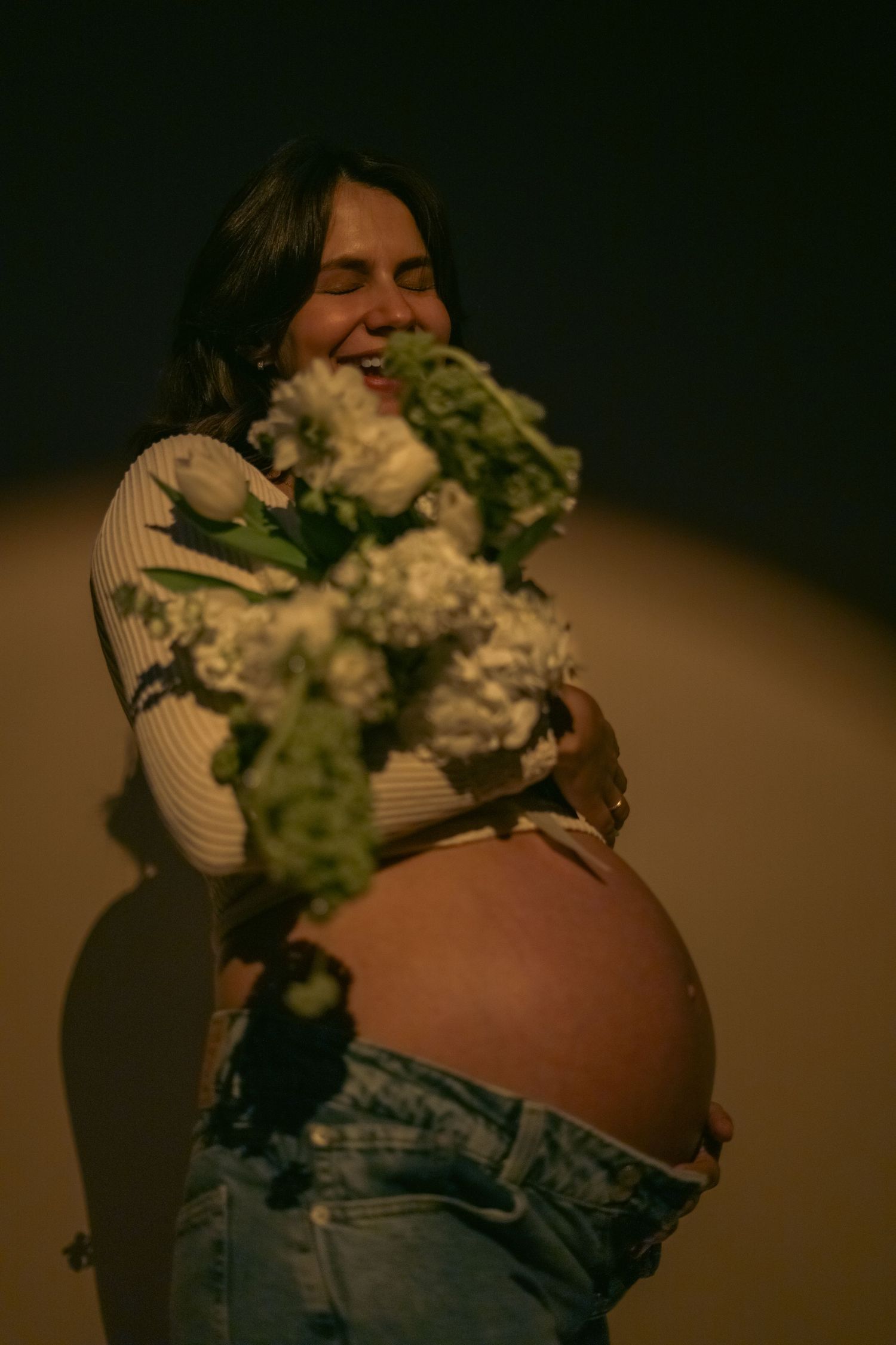 Smiling pregnant woman in jeans holding white flowers against her bare belly in warm golden light.