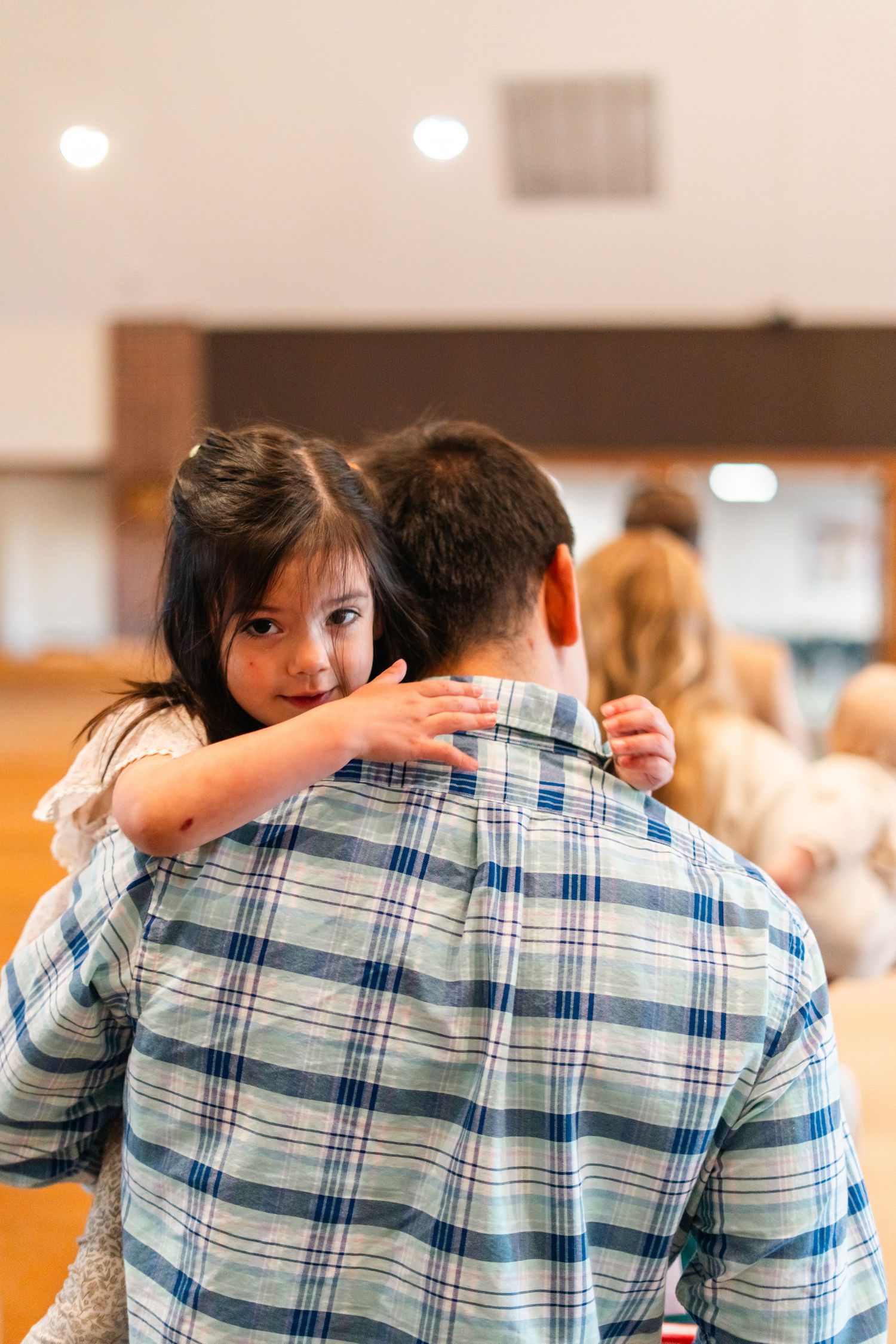 Young girl with dark hair smiles over the shoulder of a man in a plaid shirt in a bright indoor setting.