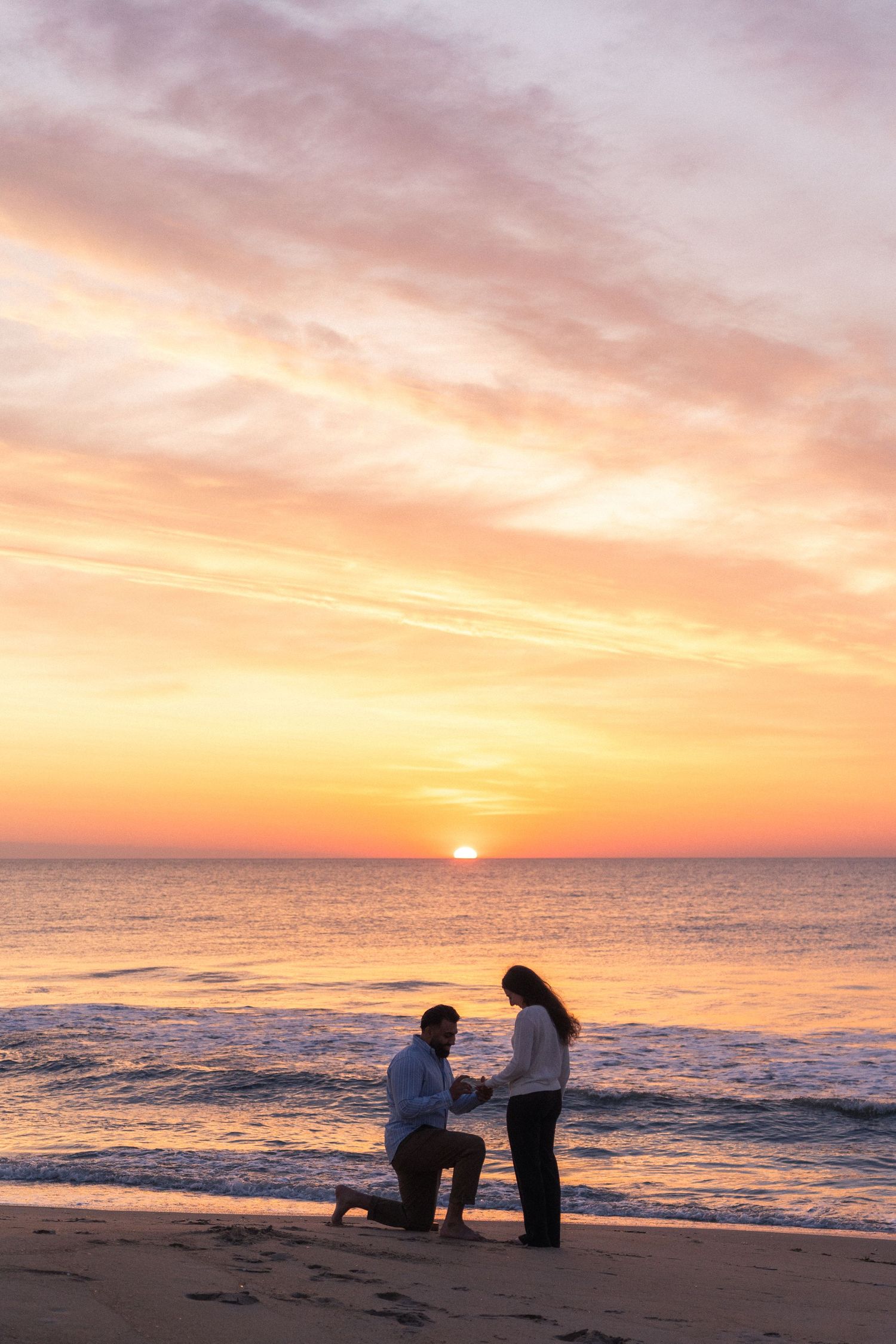 Man proposing on one knee to woman on a sandy beach at golden sunset with waves and colorful sky.