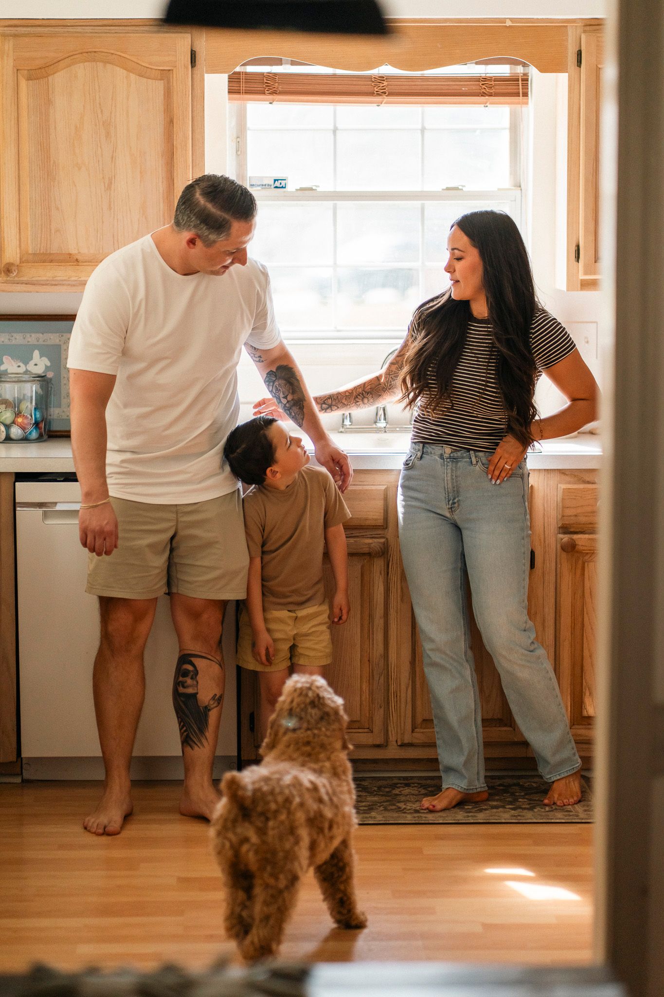 Family with young child and brown dog standing together in a warm wooden kitchen.
