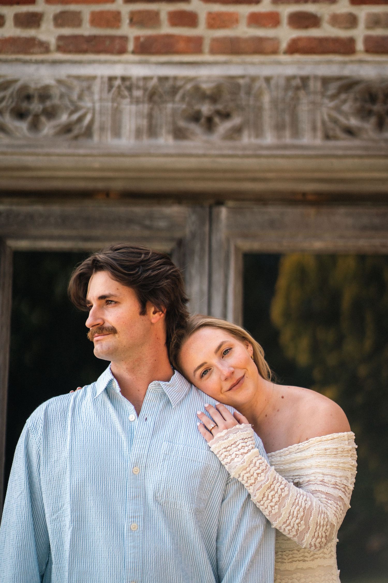 Couple posing in front of ornate brick building, man in blue shirt with woman in white lace top.