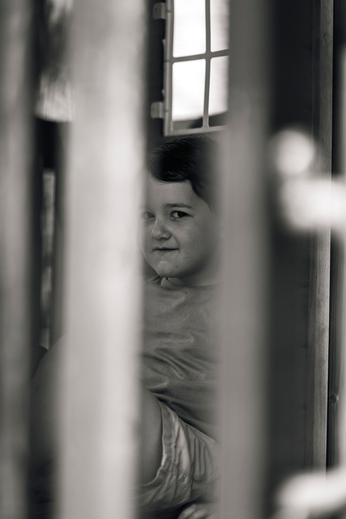 Black and white portrait of smiling baby peeking through wooden crib slats.