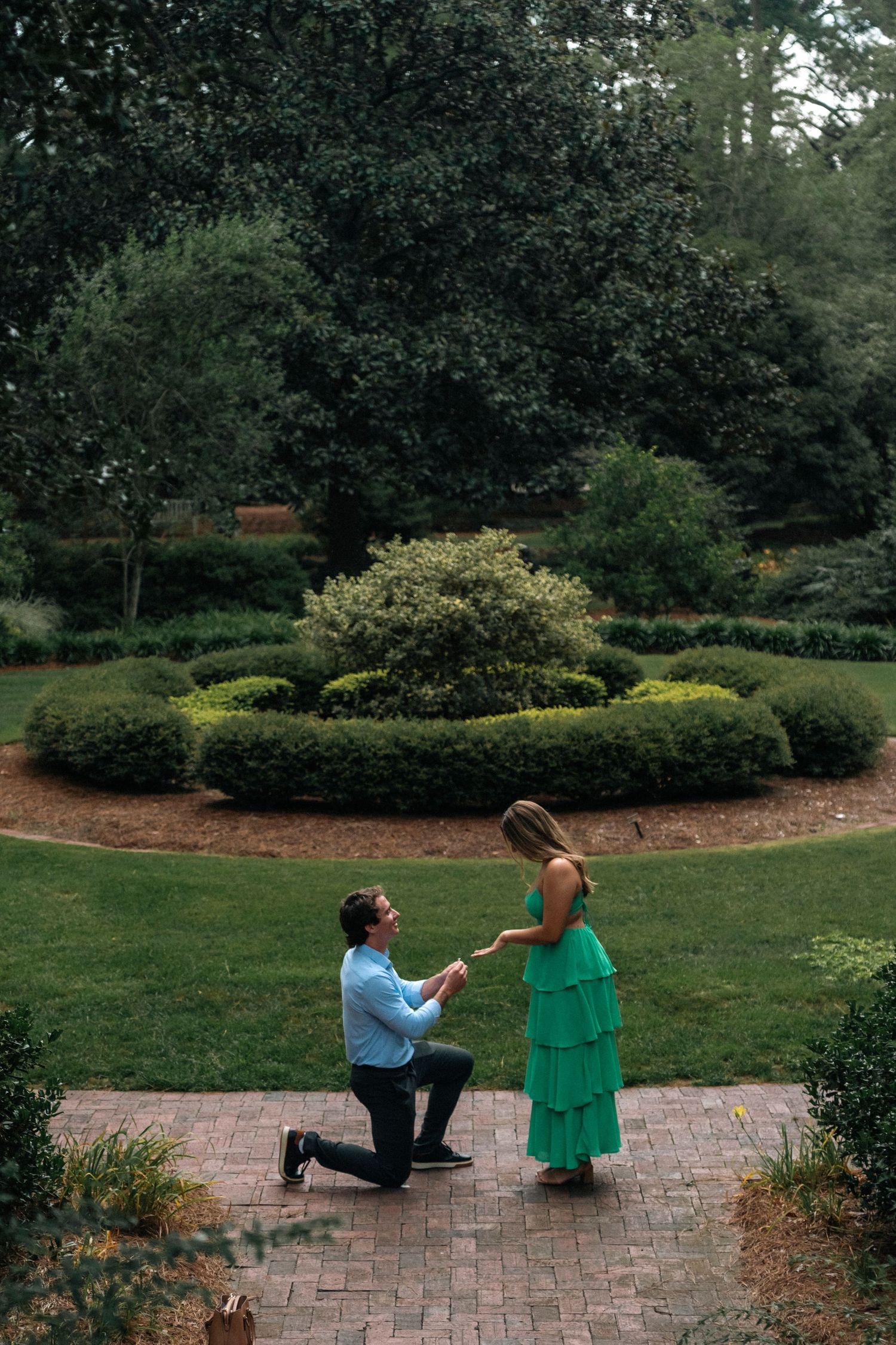 Man proposes on one knee to woman in green dress in a lush garden setting with manicured hedges.
