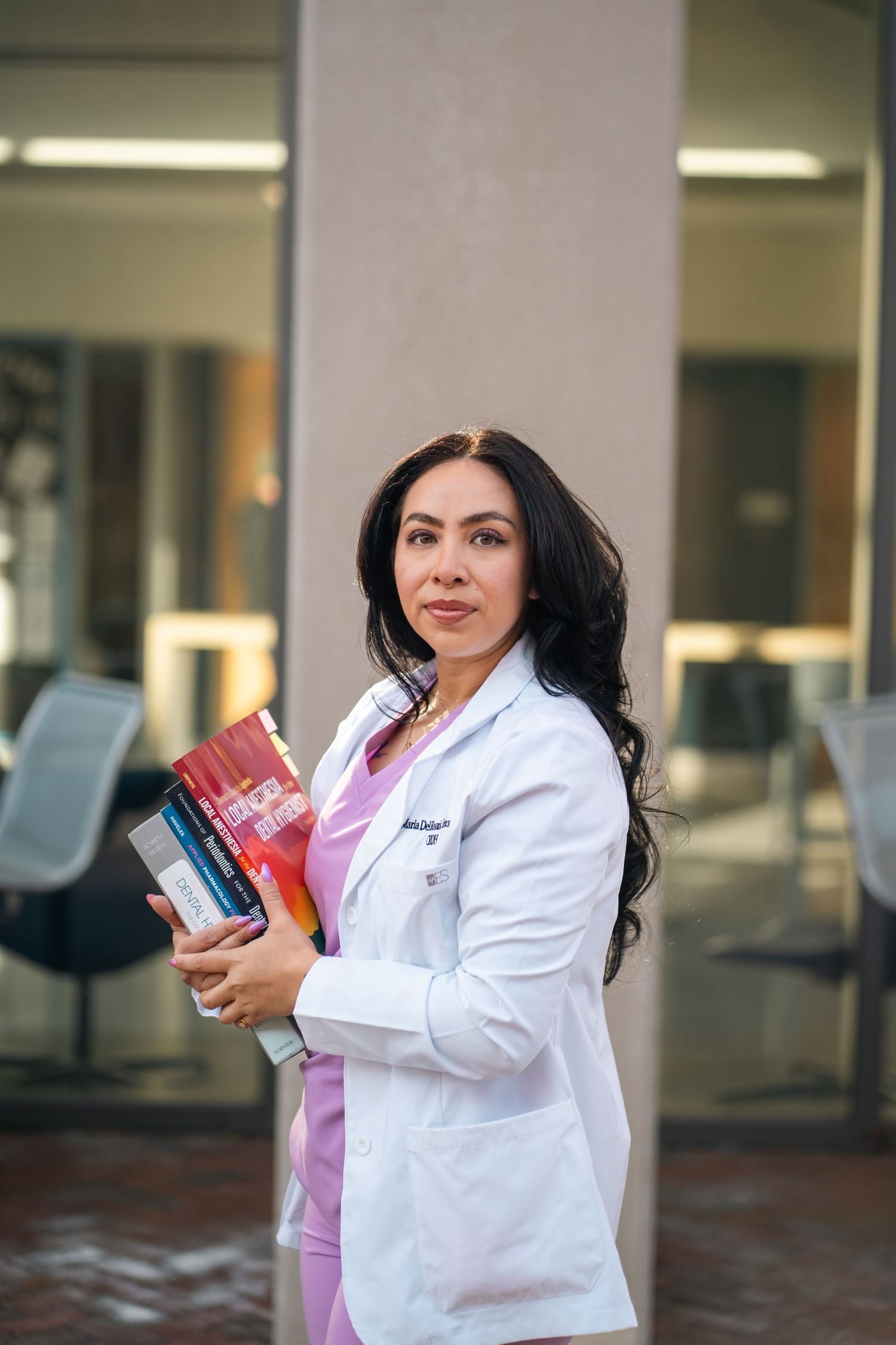 Confident female medical professional in white coat holding textbooks outside a modern building.