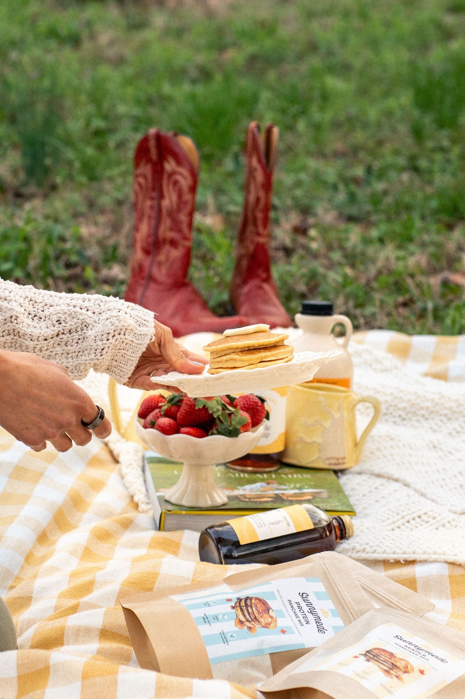 A hand reaches for crackers on a tiered stand with strawberries at a picnic with red cowboy boots in the background.