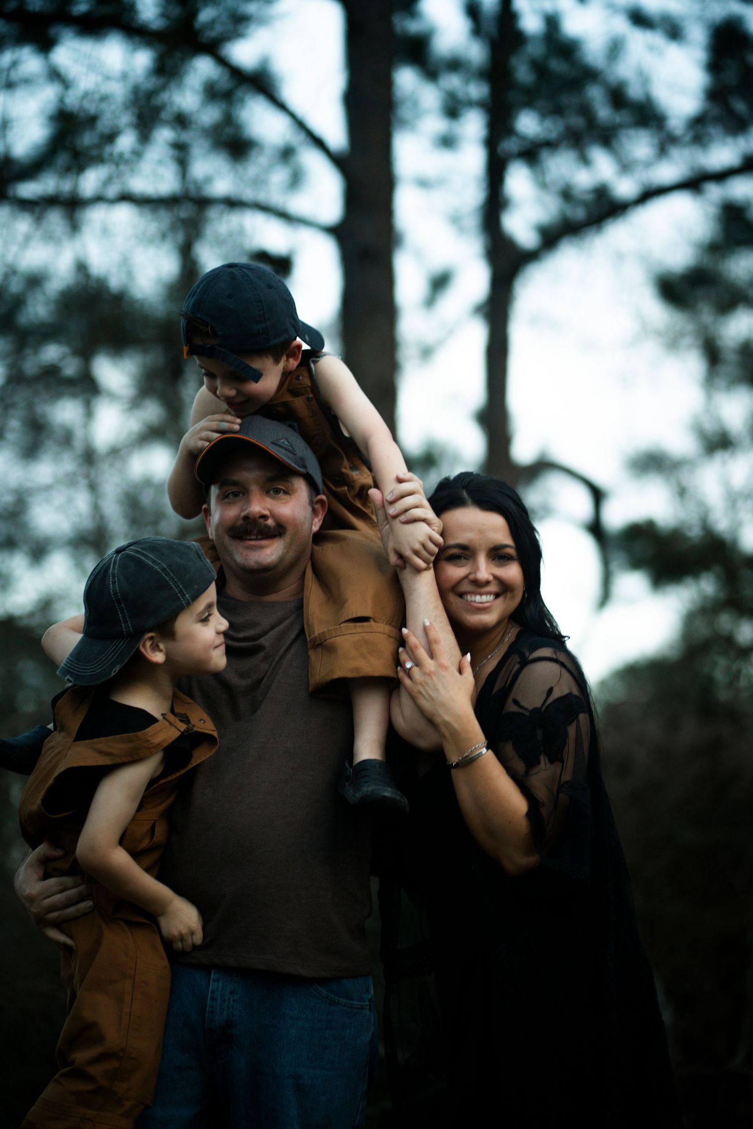 A happy family of four poses outdoors in a forest, with a child sitting on dad's shoulders, all wearing dark clothing.