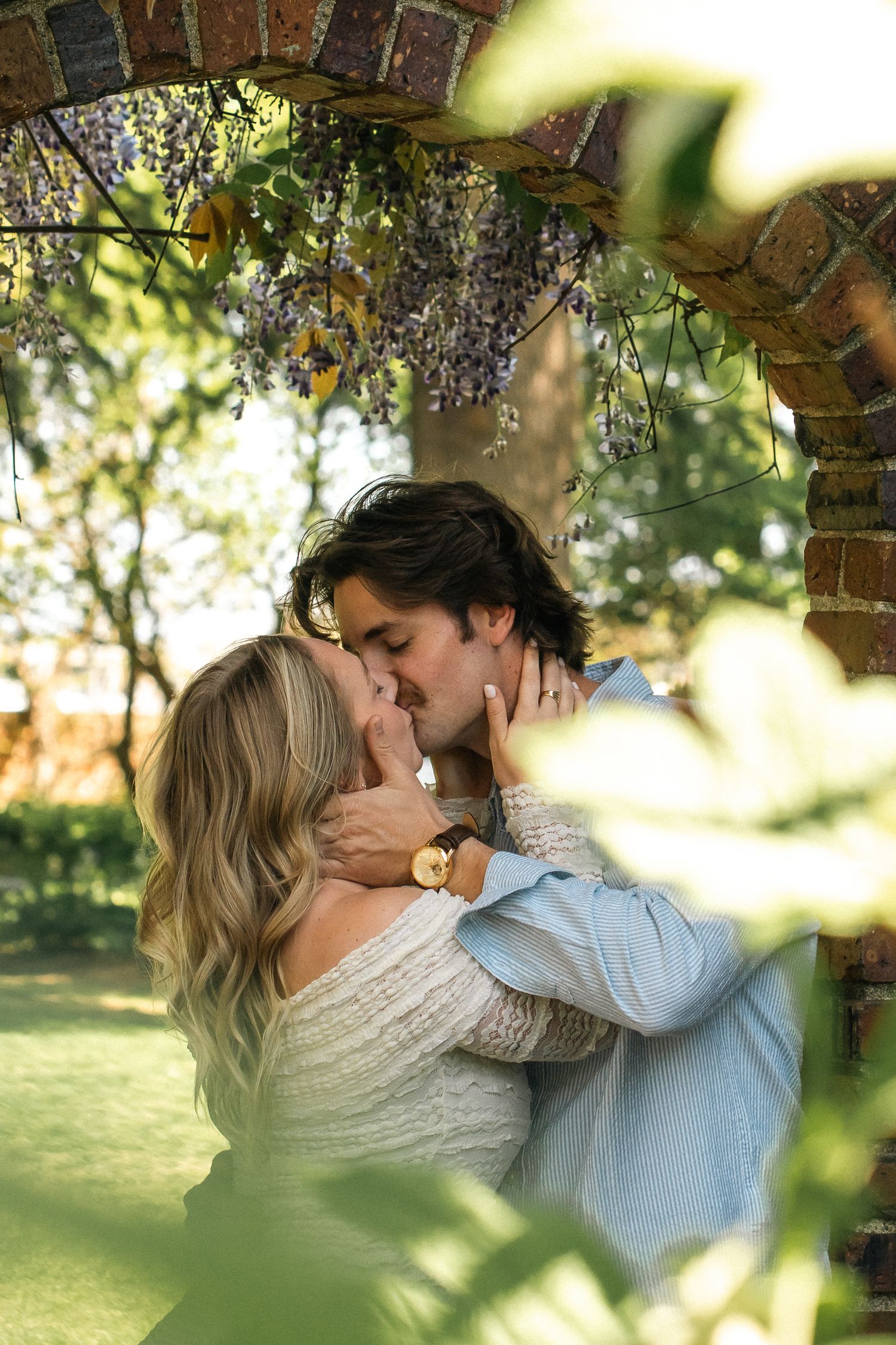 A couple shares a romantic kiss under a wisteria-covered brick arch in a sunny garden.