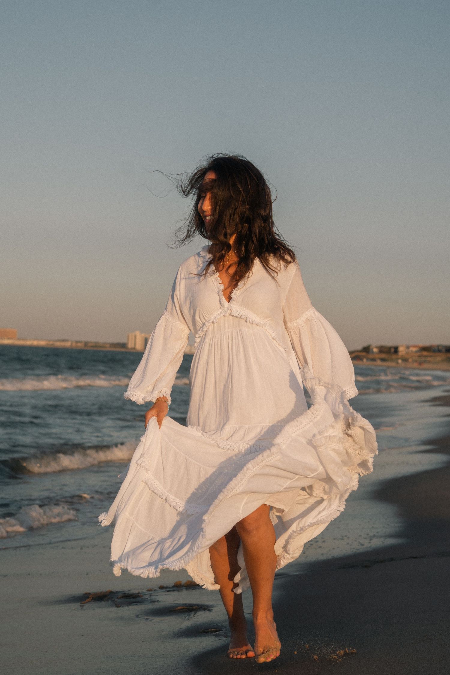 Woman in flowing white dress walking barefoot on beach with wind in her hair at sunset.
