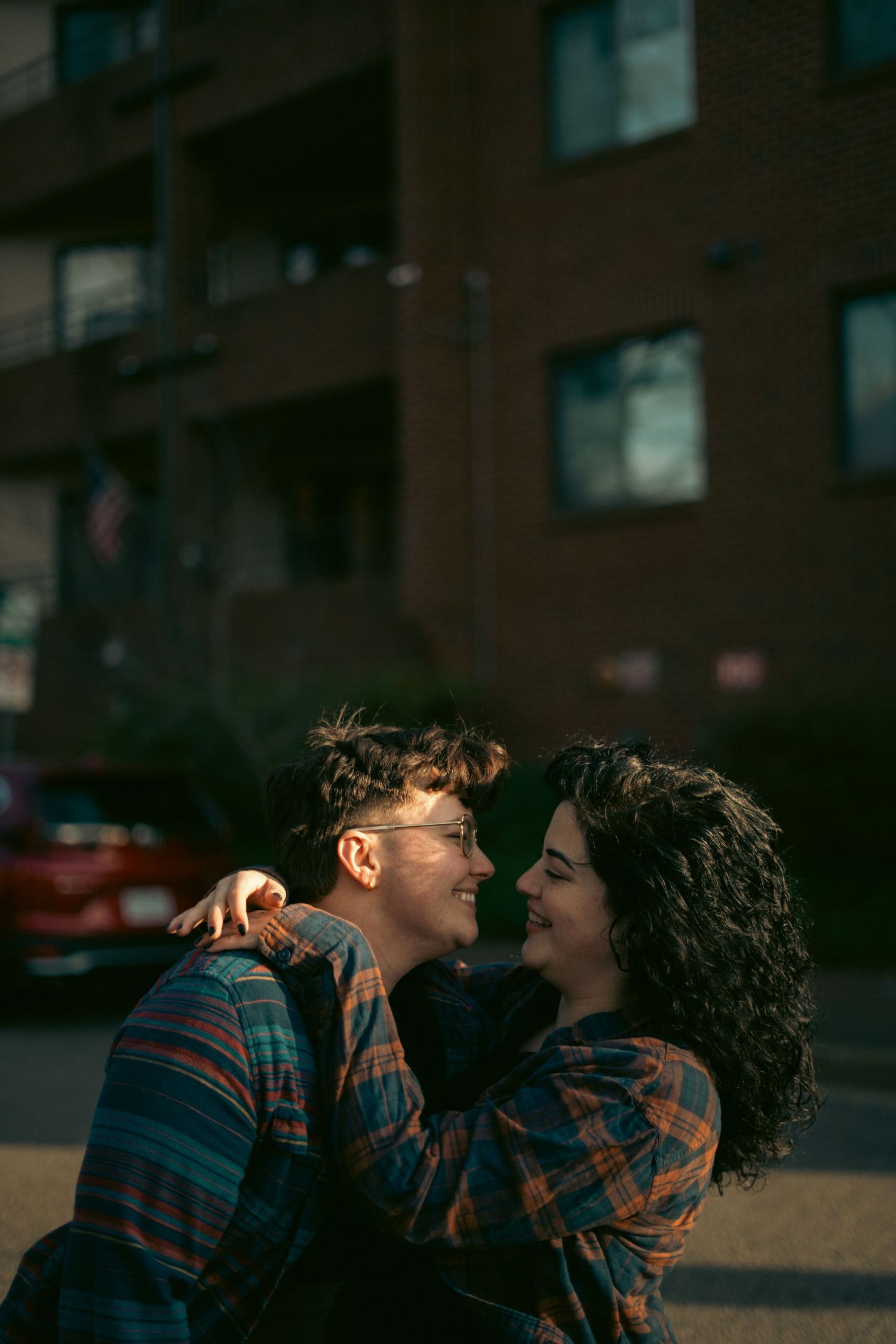 A smiling couple embraces outdoors near a brick building, sharing a joyful moment in casual plaid shirts.