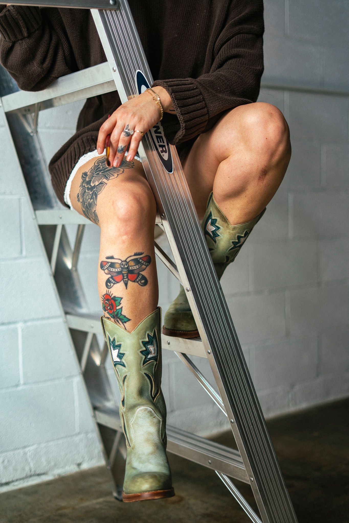 Tattooed legs wearing green cowboy boots rest on a metal ladder against a white brick wall.