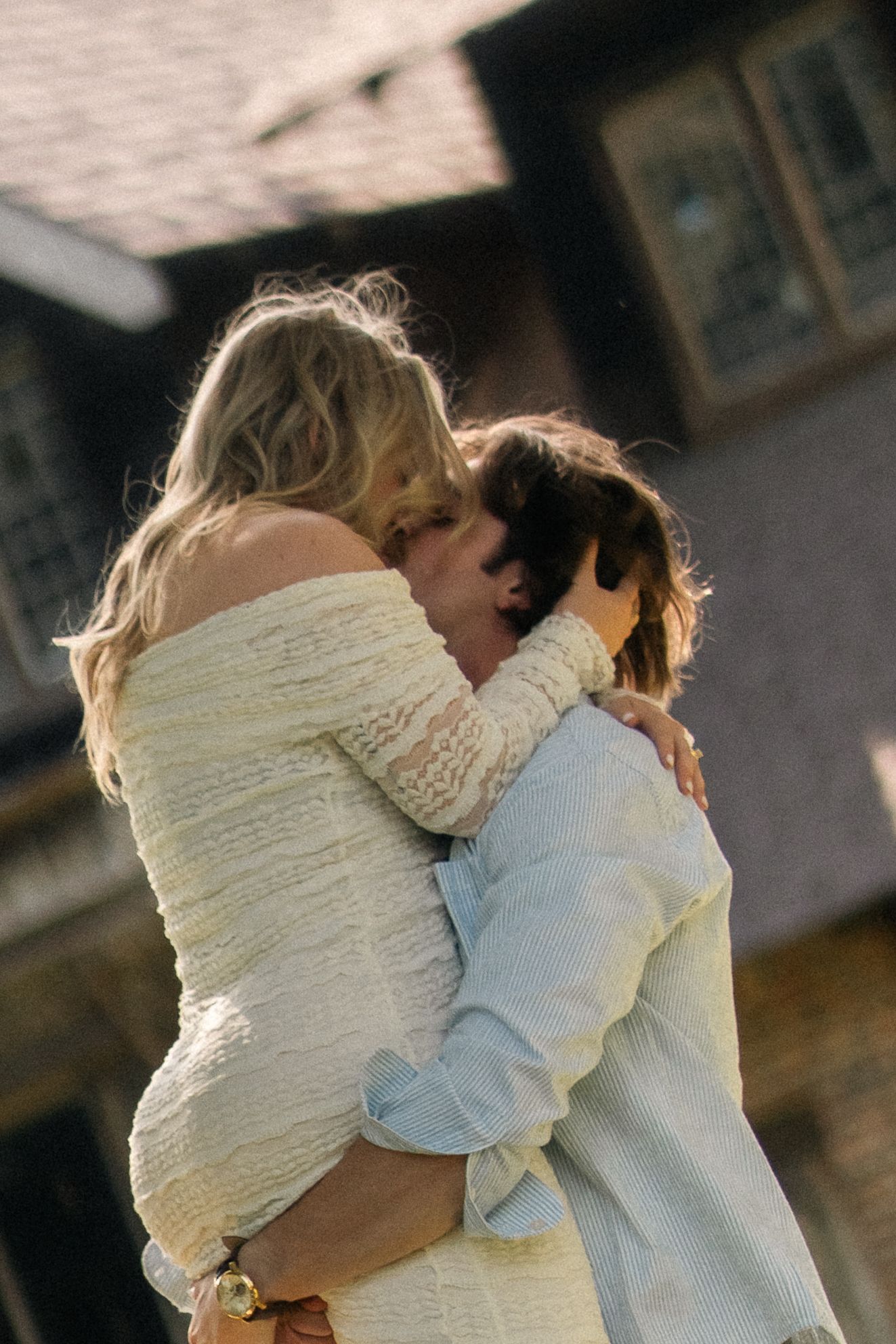 A couple embracing warmly outdoors near a building, woman in white off-shoulder dress hugging a man in a light blue shirt.