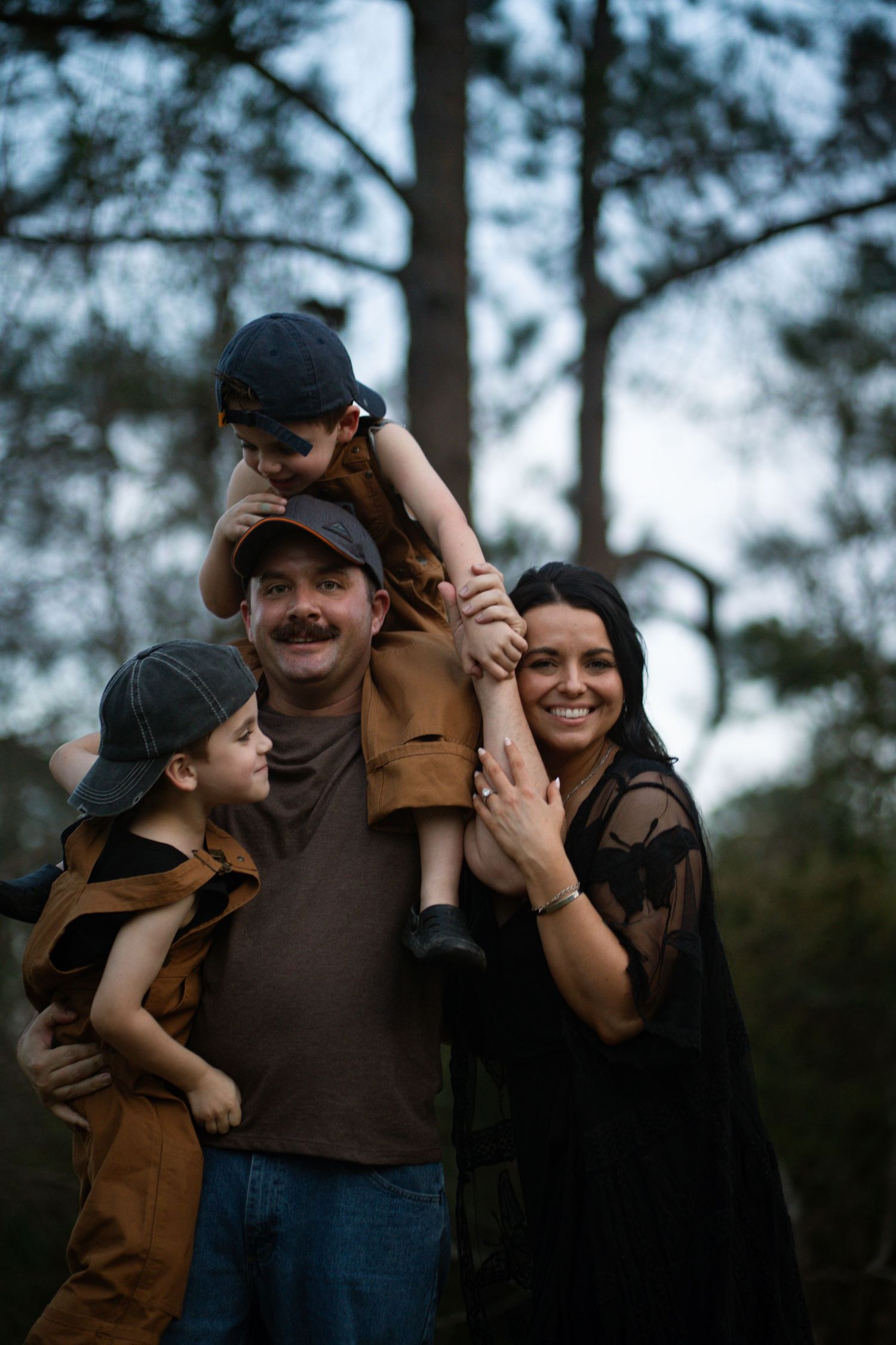 Family of twin boys, father with a mustache, and a woman wearing a lacy duster. They are all hugging, with one twin on the fathers shoulder