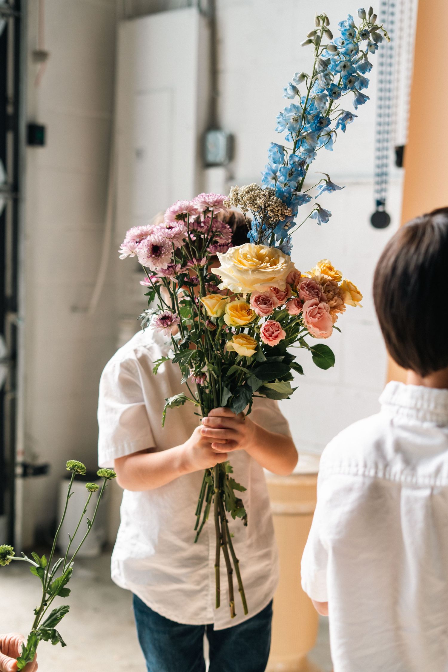Child holding large bouquet of mixed flowers including blue delphiniums and yellow roses indoors.