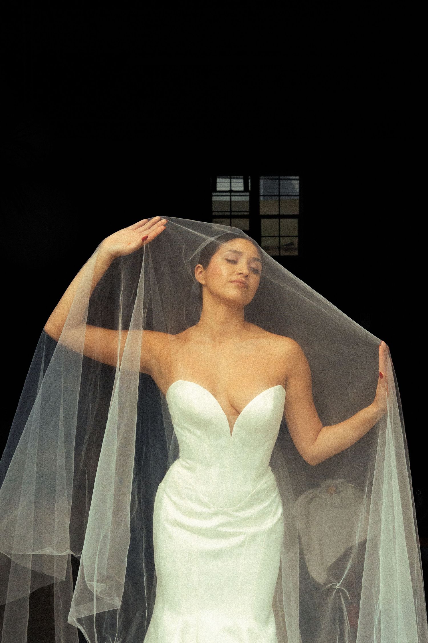 Bride in strapless sweetheart mermaid gown lifting sheer veil overhead against dramatic black background.
