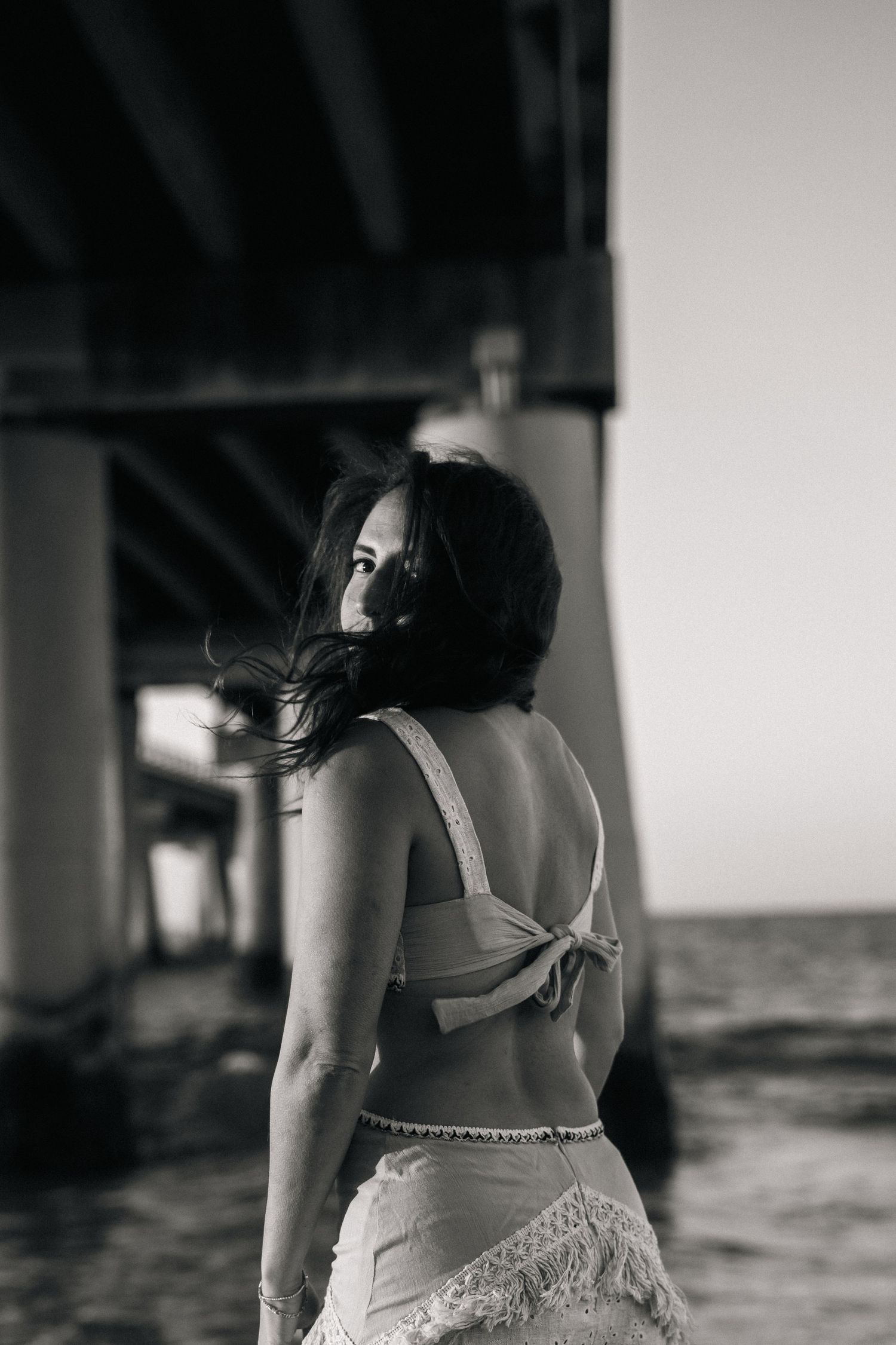 Black and white photo of woman in white bikini top standing under pier by the ocean.
