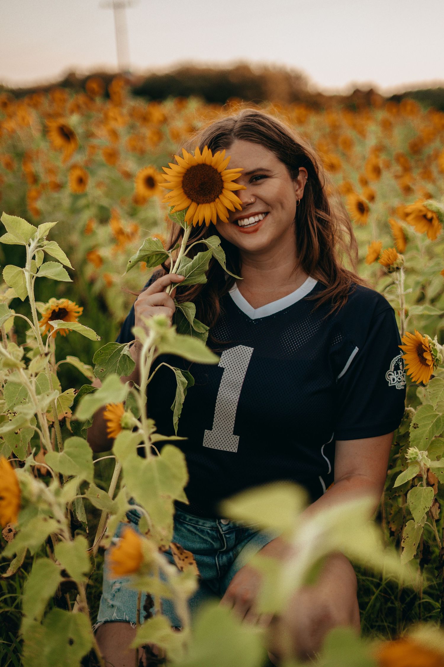 Smiling woman in football jersey holding sunflower over eye while sitting in sunflower field at sunset.