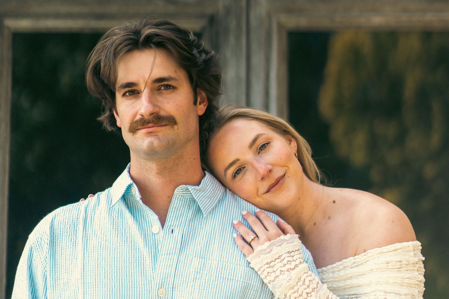Romantic couple portrait with woman leaning on man's shoulder, both smiling softly outdoors in natural light.