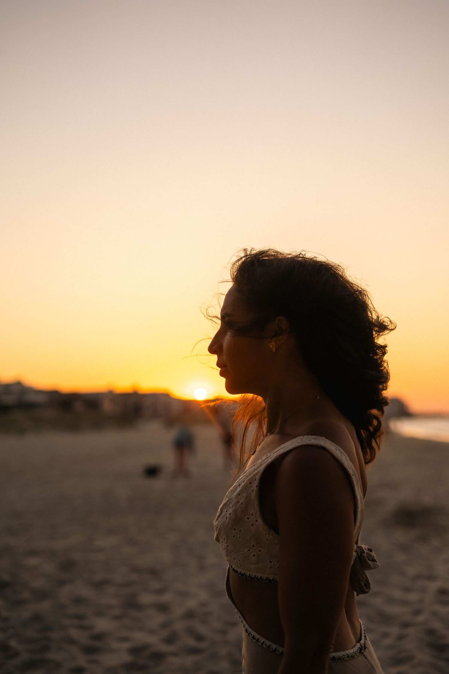 Side profile of woman silhouetted against warm orange sunset sky on sandy beach.