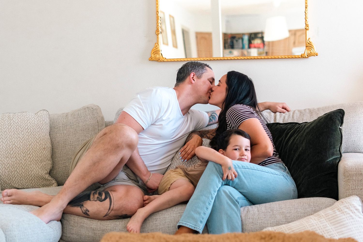 Happy family with parents kissing and young child smiling while relaxing together on a cozy living room sofa.