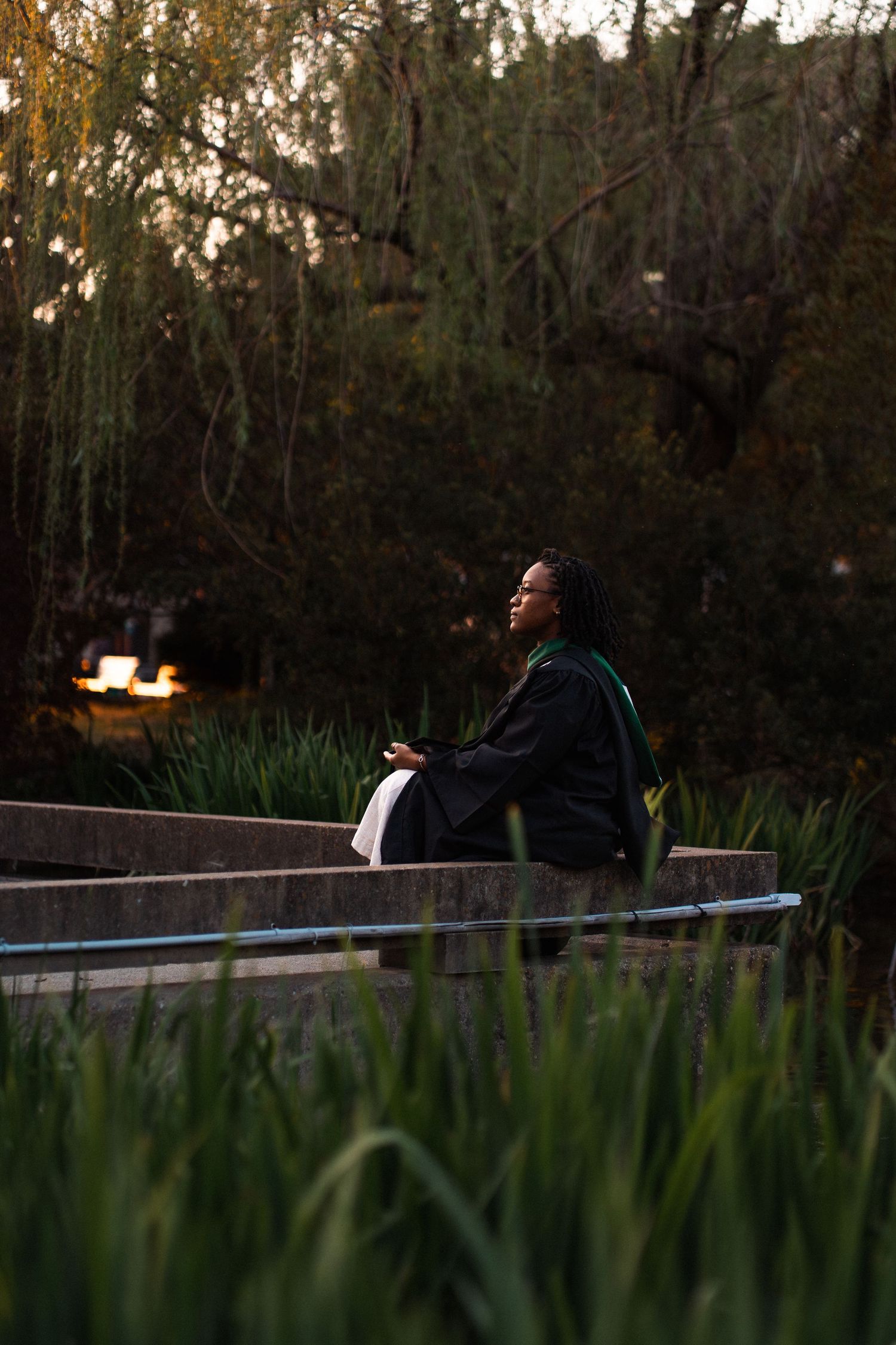 A graduate in cap and gown sits peacefully on stone steps surrounded by lush green trees at dusk.