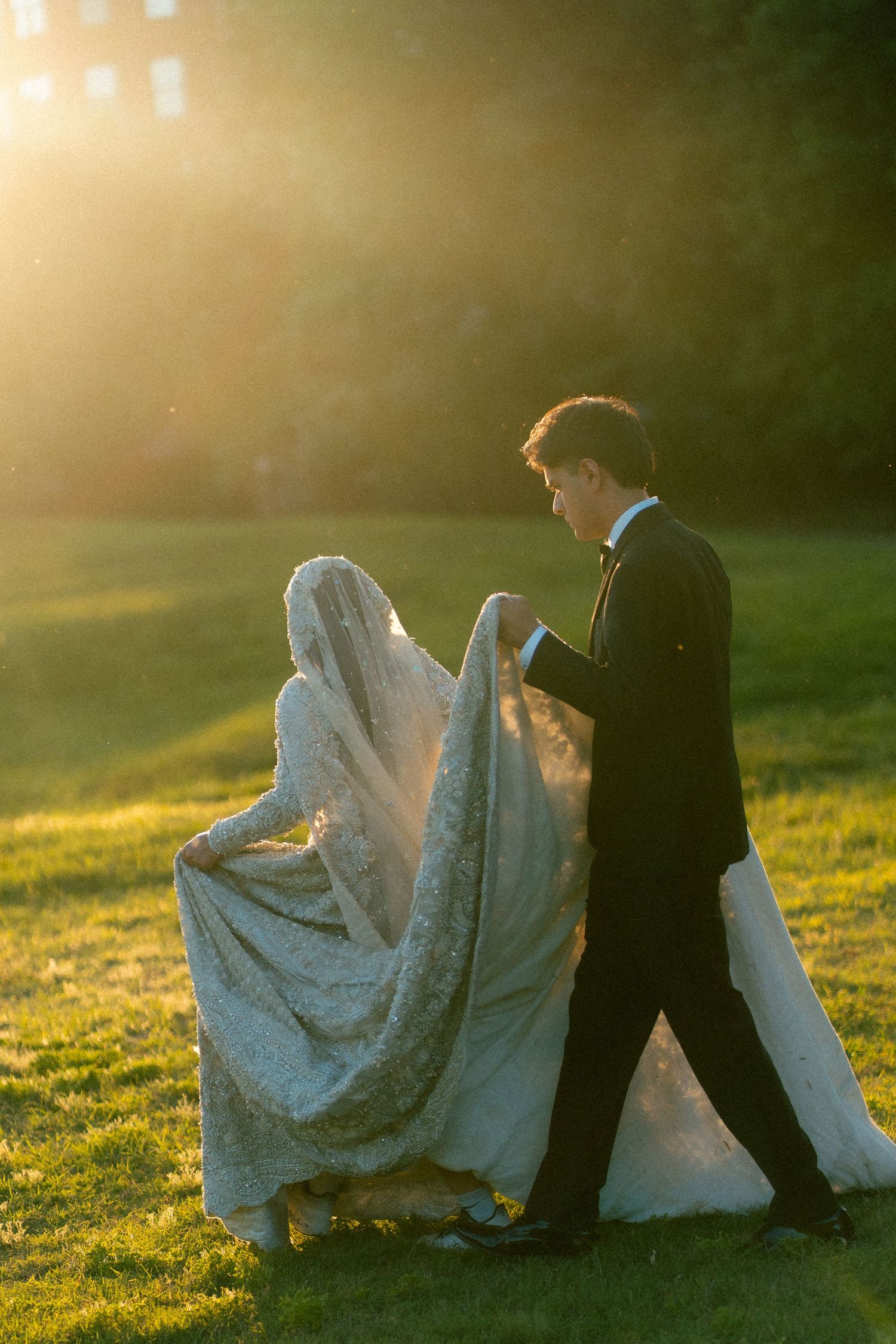 Bride and groom walking on green grass at golden hour sunset, bride in lace gown with long veil, groom in black tuxedo.