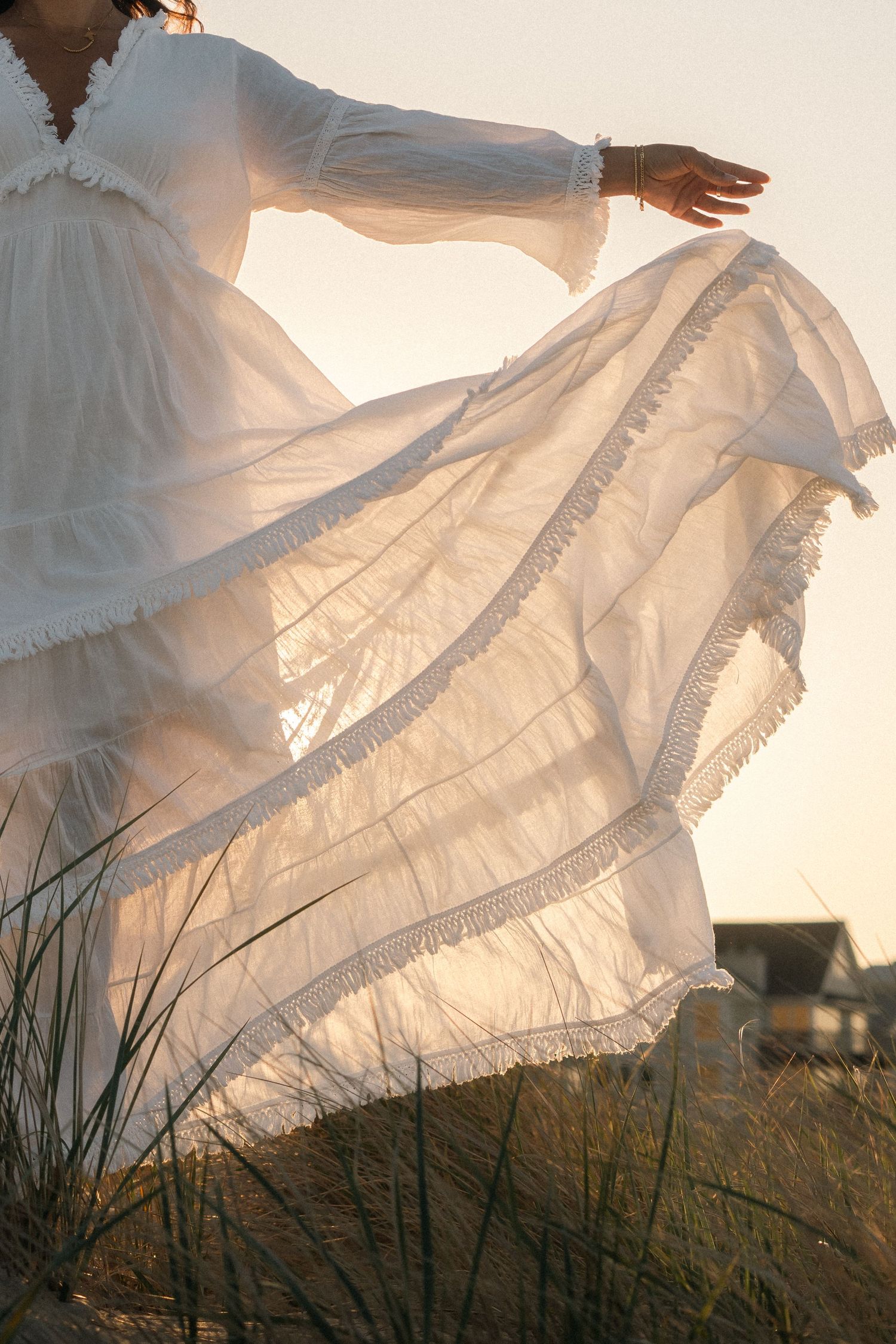 Woman in flowing white boho dress with fringe twirling on sandy dunes at sunset.