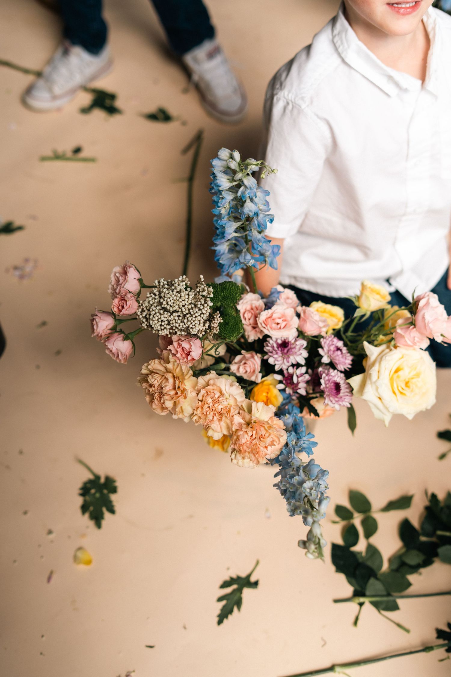 A smiling child in a white shirt holds a large colorful bouquet of mixed flowers on a beige floor.